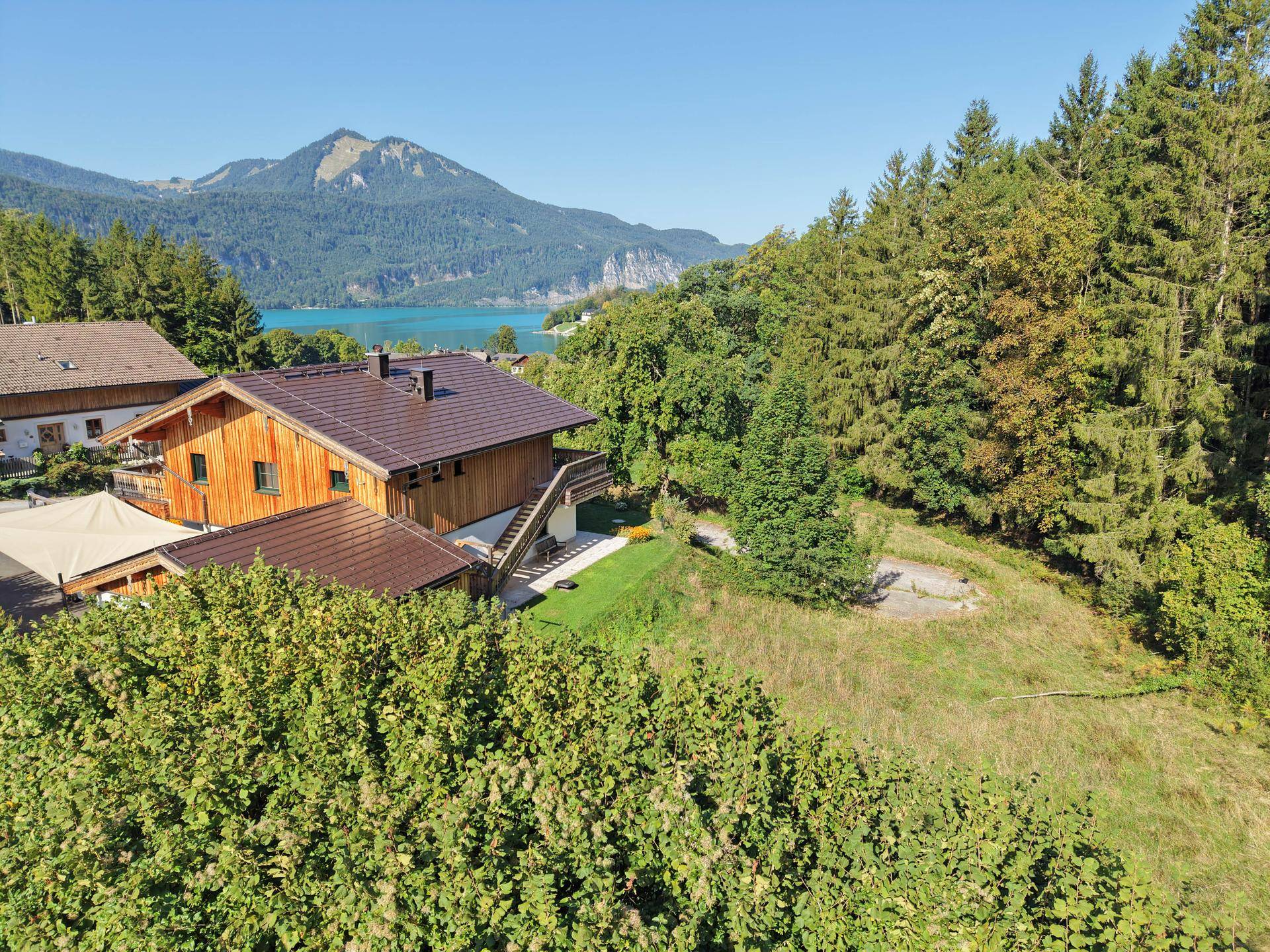 Panoramablick auf ein Holzhaus mit weitläufigem Grundstück, Wald und einem See im Hintergrund.
