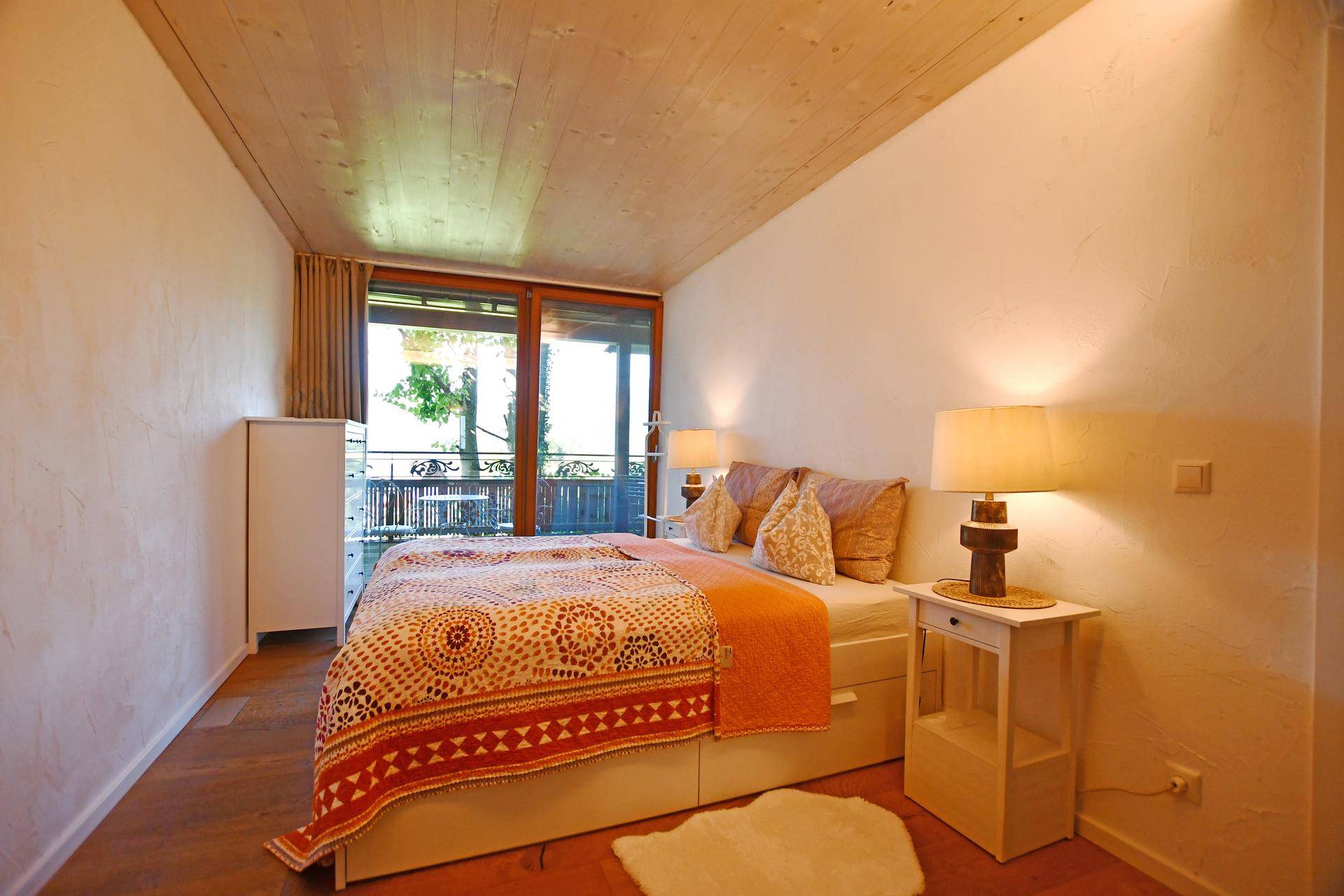 Helles Schlafzimmer mit Doppelbett, Holzboden und Zugang zu einem Balkon oder einer Terrasse.