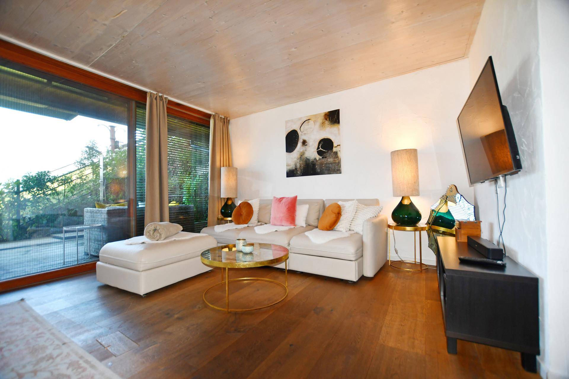 Gemütliches Wohnzimmer mit beigem Sofa, Holzboden und großen Fenstern zum Außenbereich.