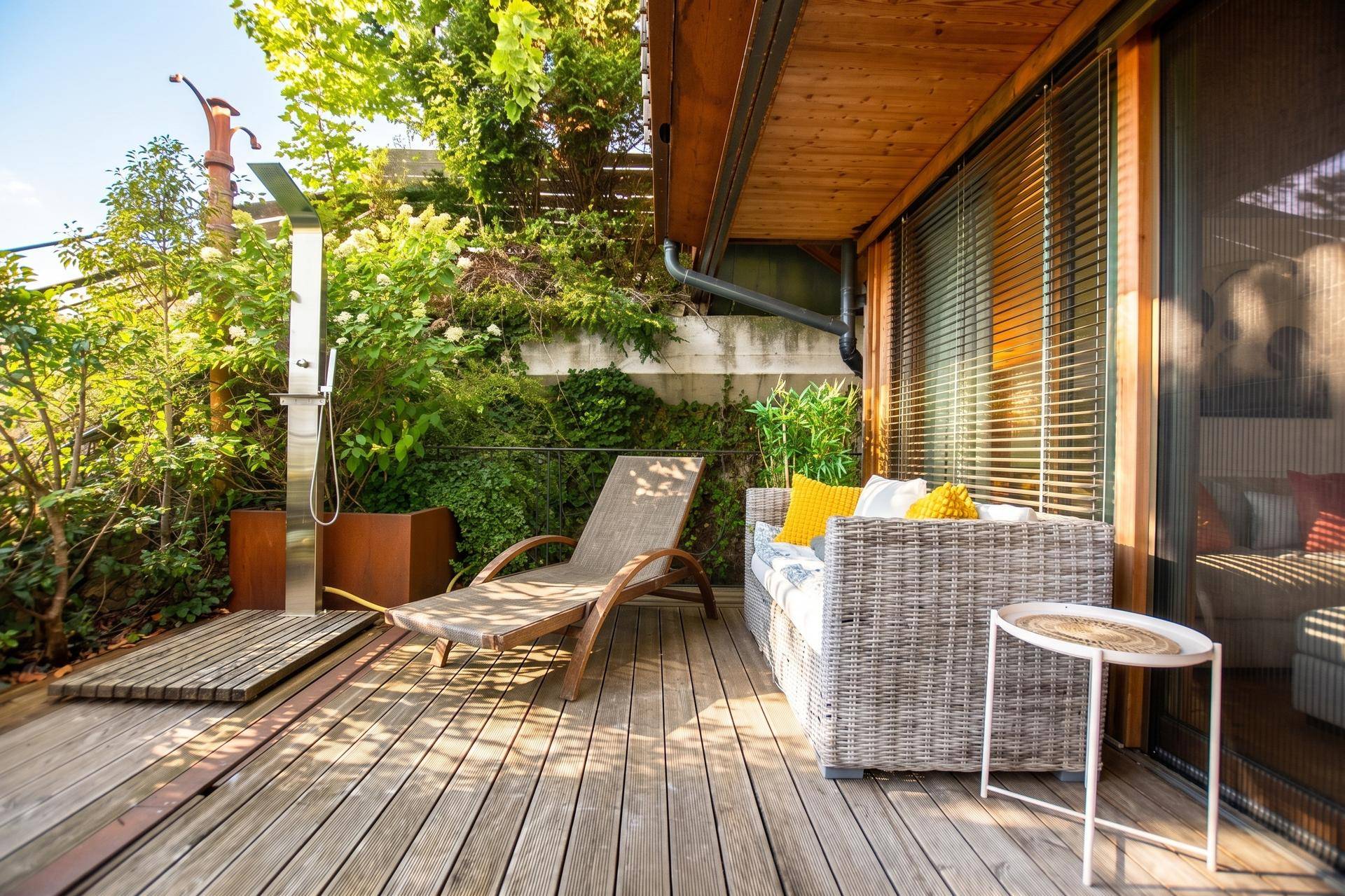 Sonniger Terrassenbereich mit Holzdeck, Liegestuhl und Outdoor-Dusche, umgeben von üppigem Grün.