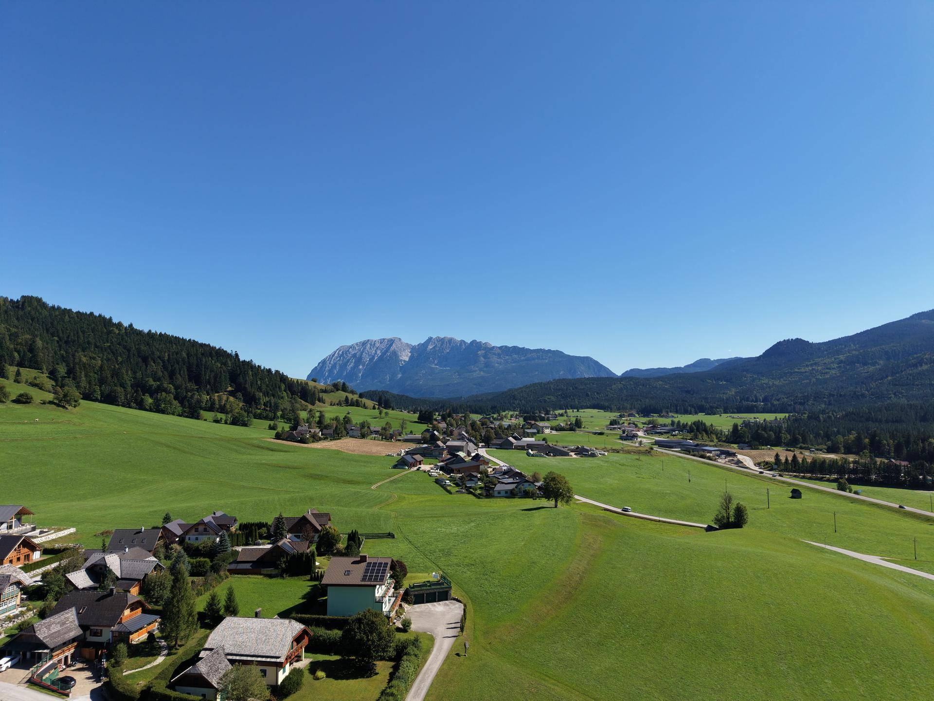 Weitläufige Landschaft mit einem Dorf, grünen Feldern und Bergen unter strahlend blauem Himmel.