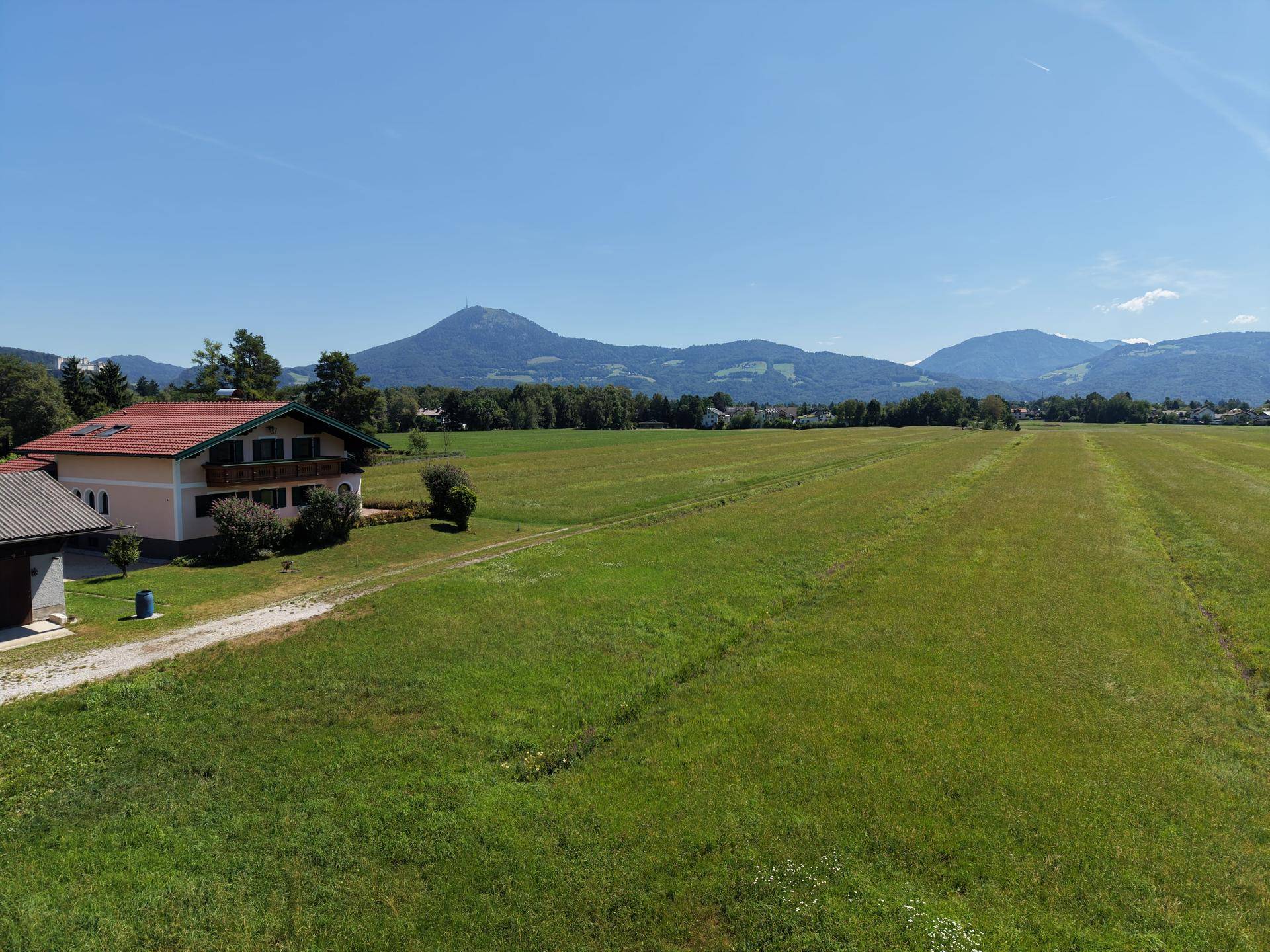 Luftaufnahme eines Hauses mit rotem Dach und weiten grünen Feldern im Vordergrund der Berge.