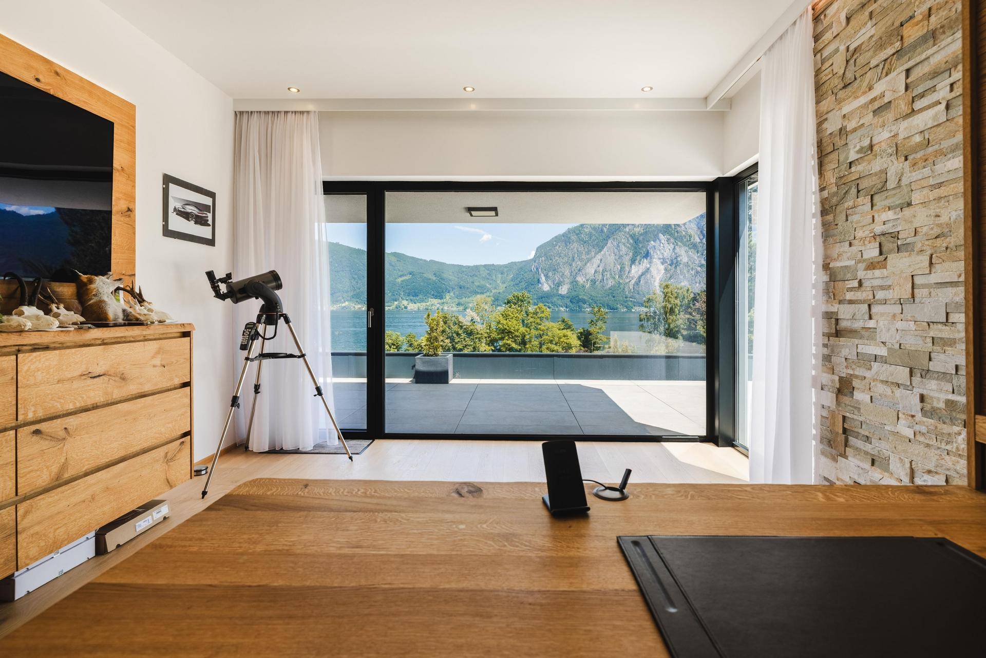 Helles Arbeitszimmer mit Holzboden, Steinwand und großem Fenster mit Panoramablick auf See und Berge.