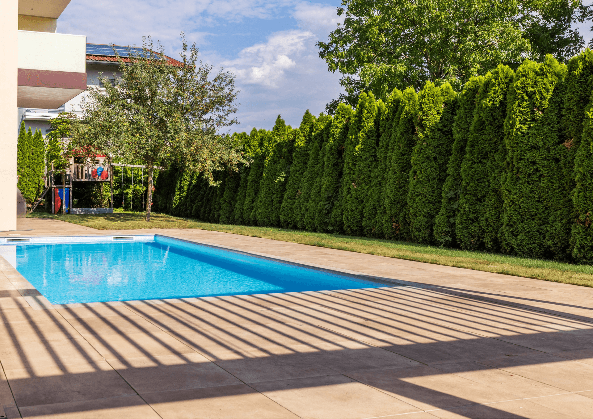 Langer Pool im Garten mit gepflegter Hecke und Kinderspielplatz im Hintergrund.