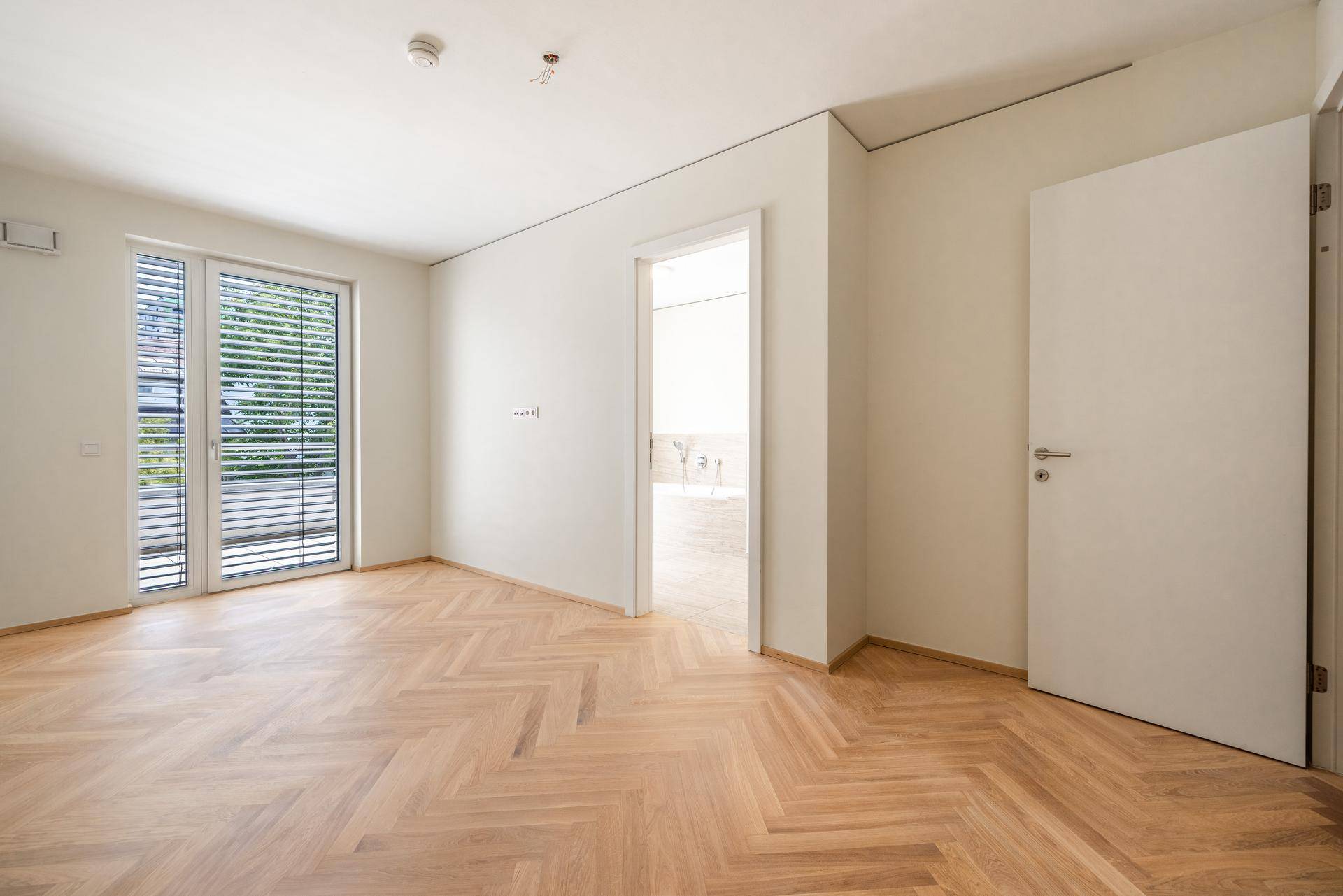 Leeres Zimmer mit Fischgrätparkett, Fenster mit Jalousien und Zugang zu einem weiteren Raum.