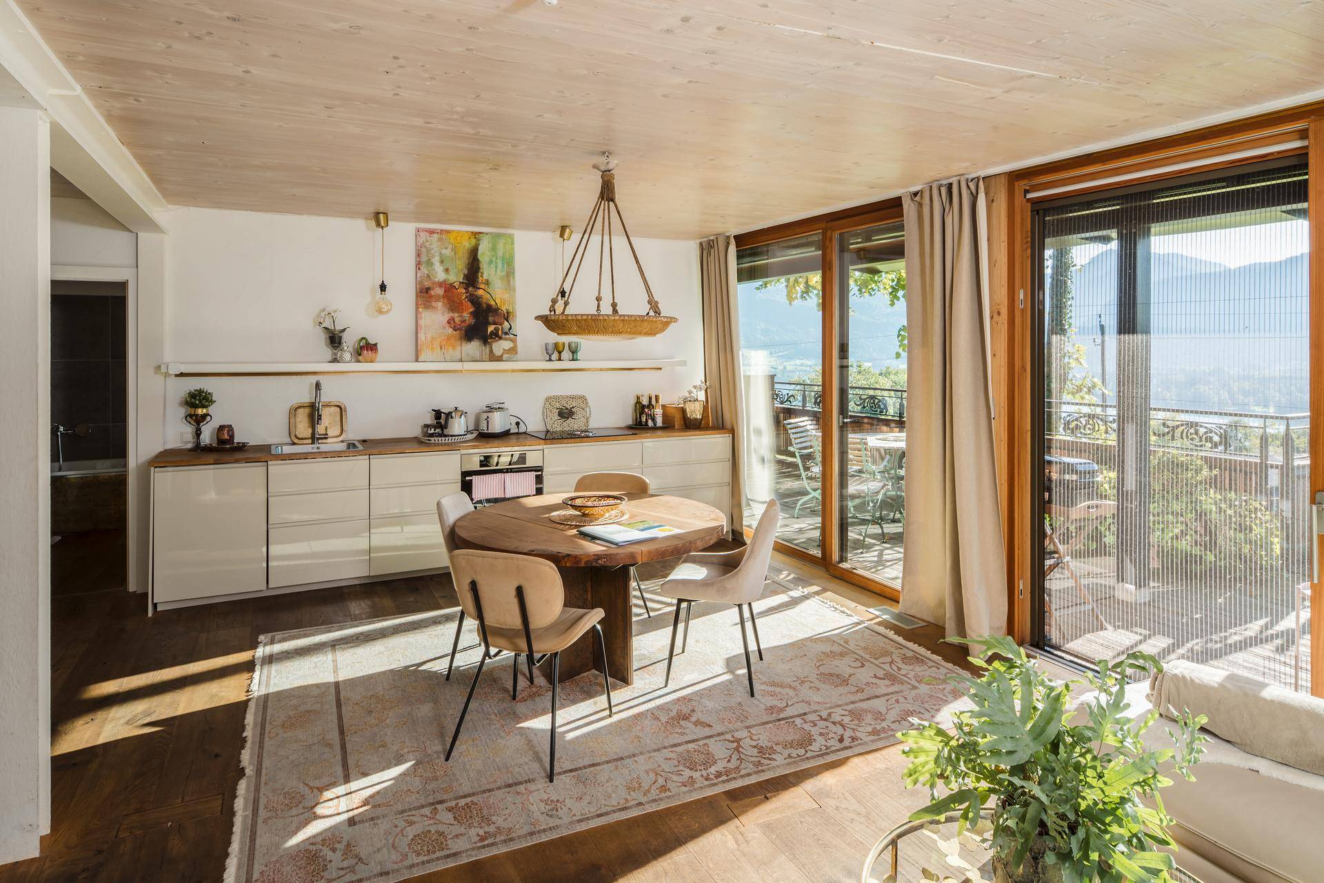Heller Wohn-Essbereich mit Holztisch, offener Küche und Zugang zu einer Terrasse mit Bergblick.