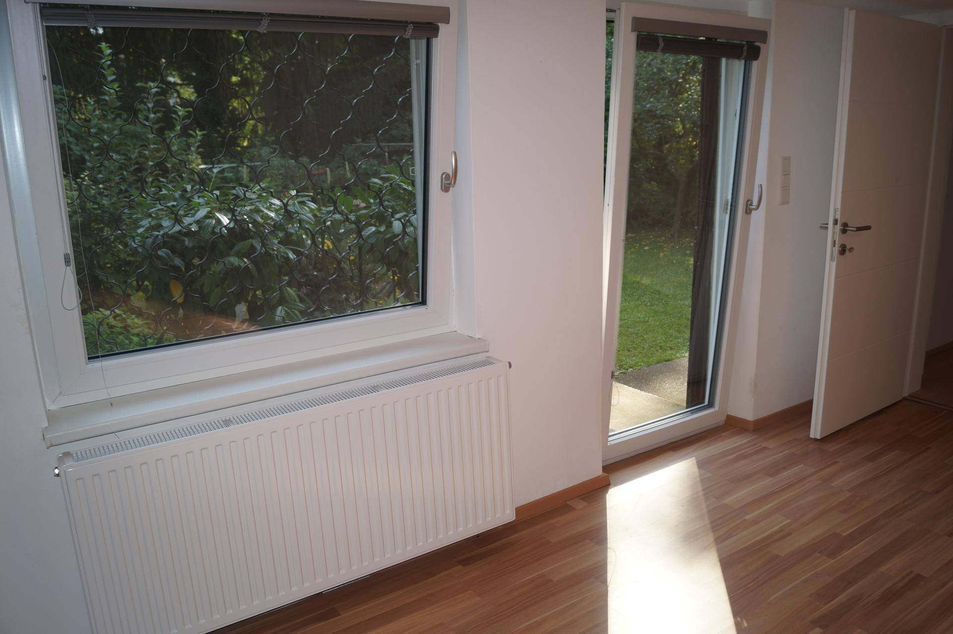 Heller Raum mit Laminatboden, Fenster und Zugang zu einem grünen Außenbereich.