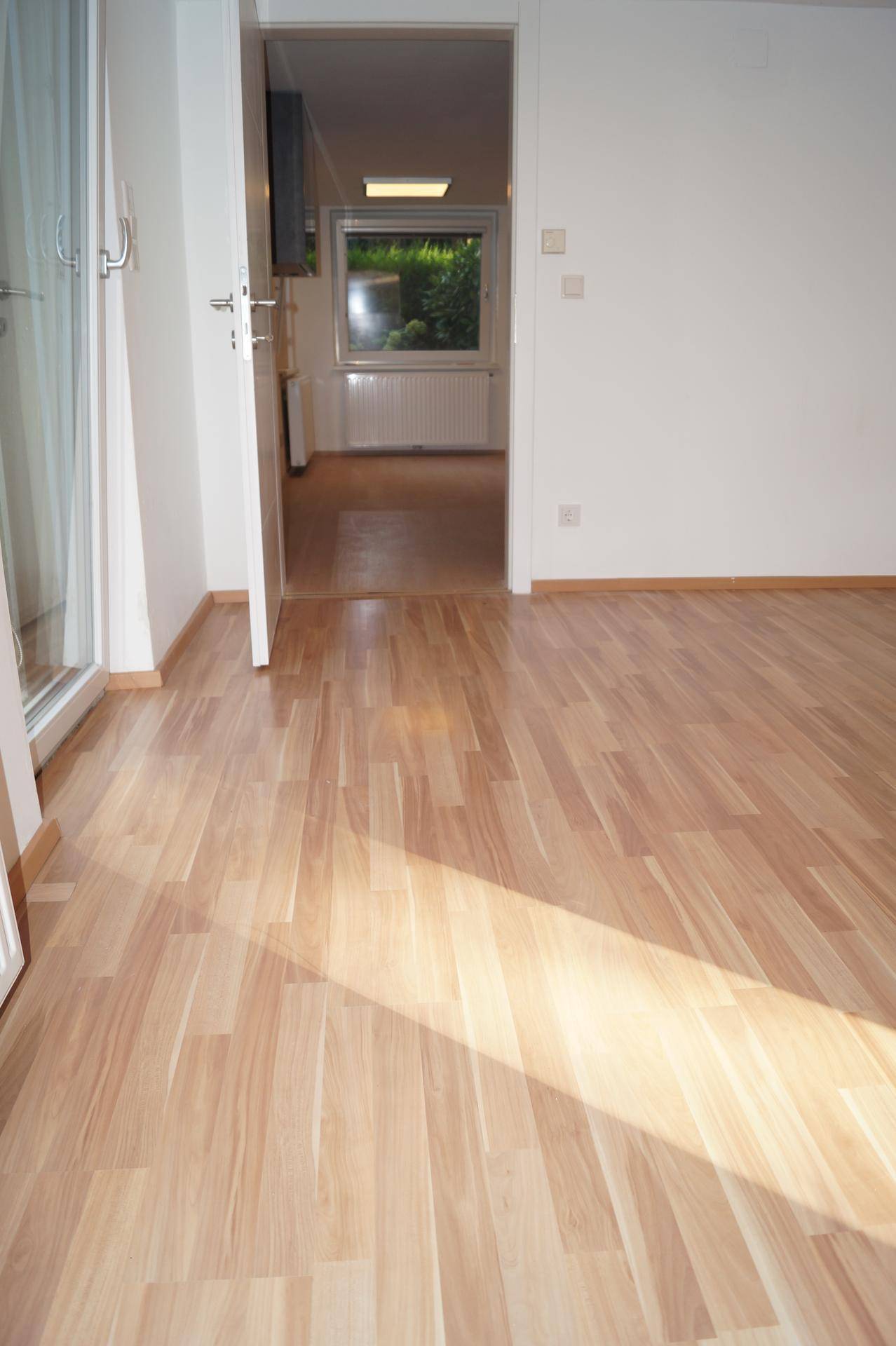 Heller Raum mit Laminatboden und Blick durch eine offene Tür in einen weiteren sonnigen Bereich.