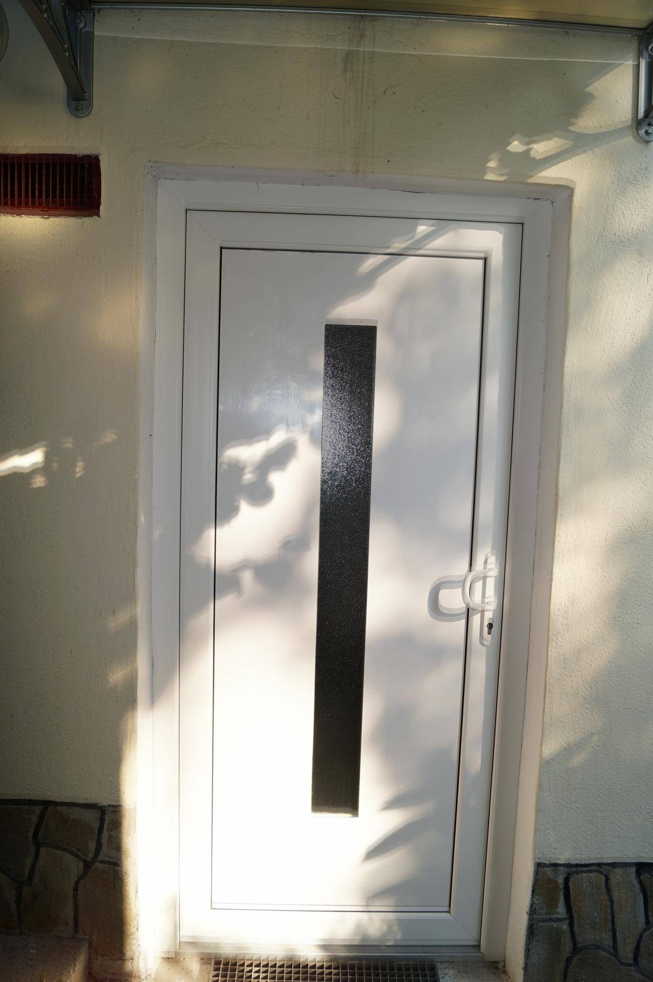 Weiße Eingangstür mit vertikalem Glaseinsatz in einer hellen Fassade, beleuchtet von der Sonne.