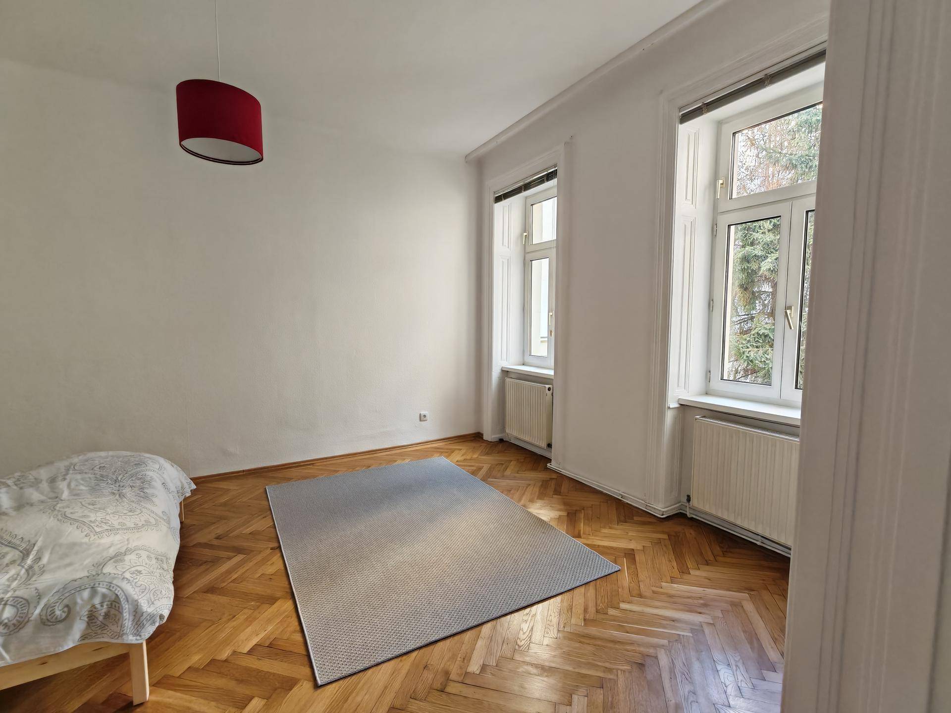 Helles Schlafzimmer mit Fischgrätparkett, zwei großen Fenstern und einer roten Deckenleuchte.