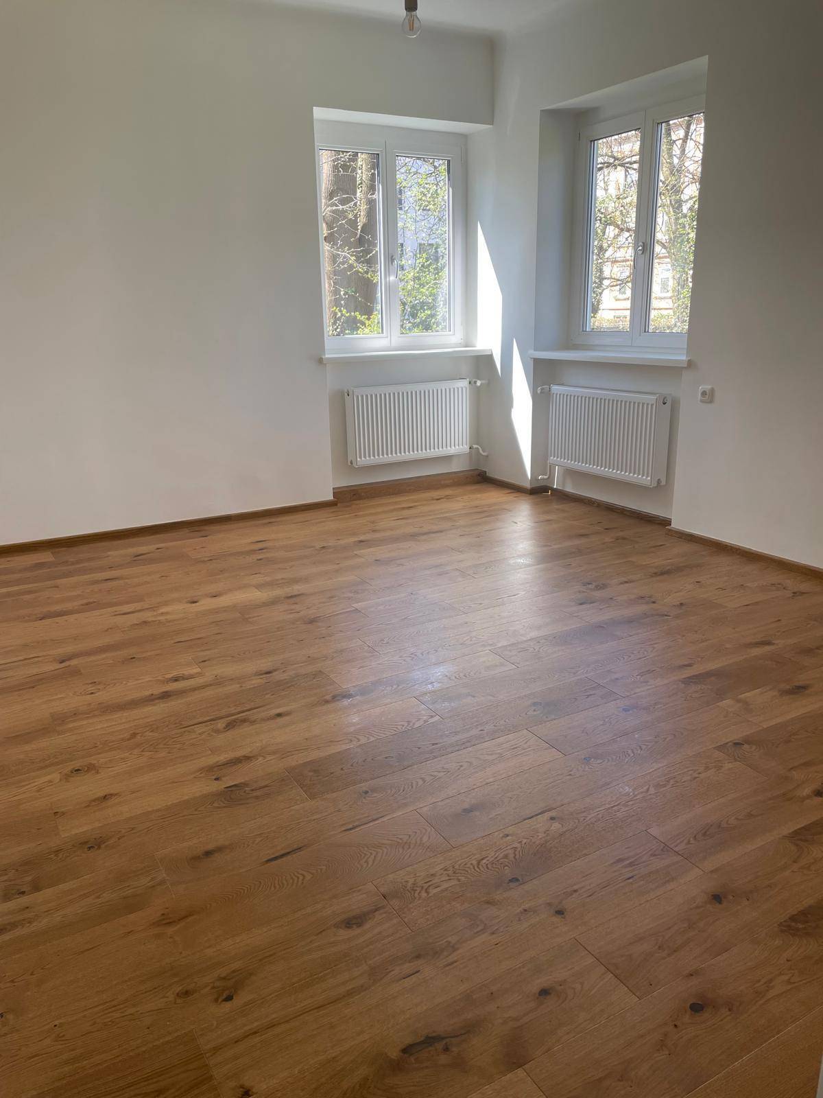 Heller, leerer Raum mit neuem Holzdielenboden und zwei Fenstern, die viel natürliches Licht hereinlassen.
