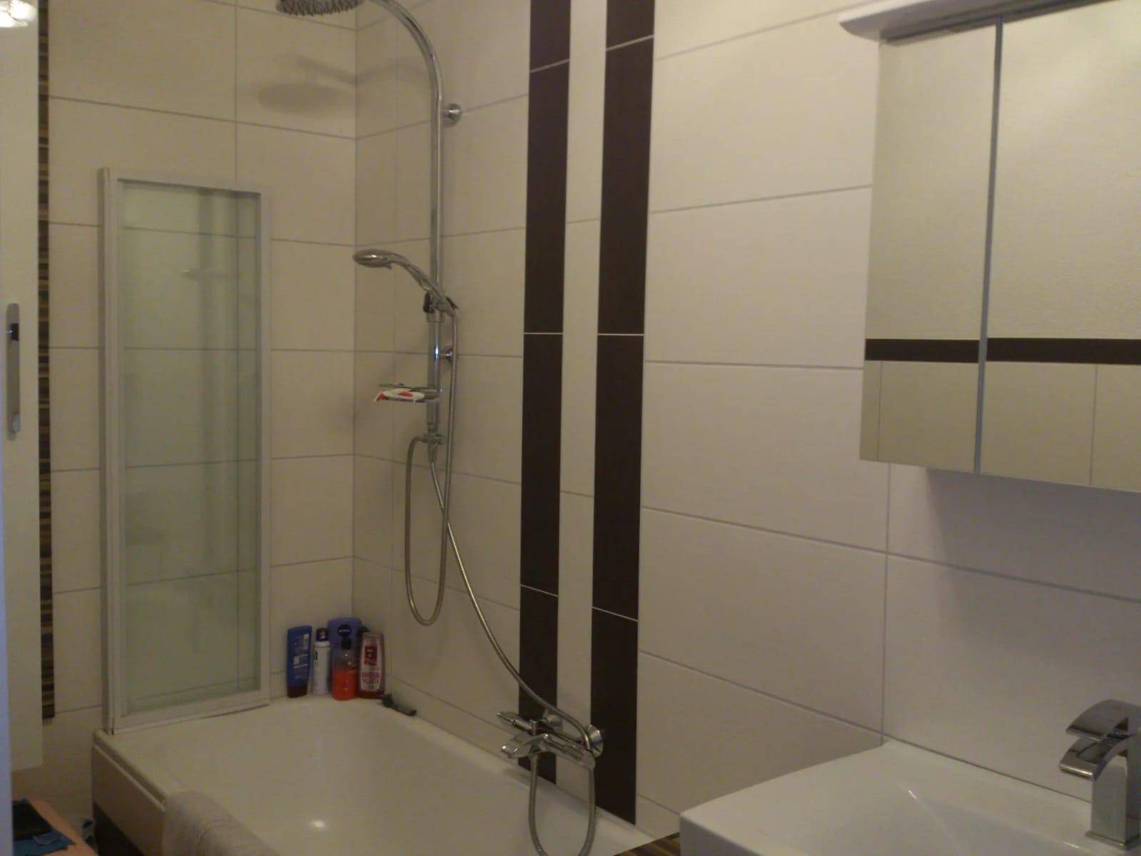 Badezimmer mit Badewanne, Duschvorrichtung und Fliesen in neutralen Farben mit vertikalen Streifen.