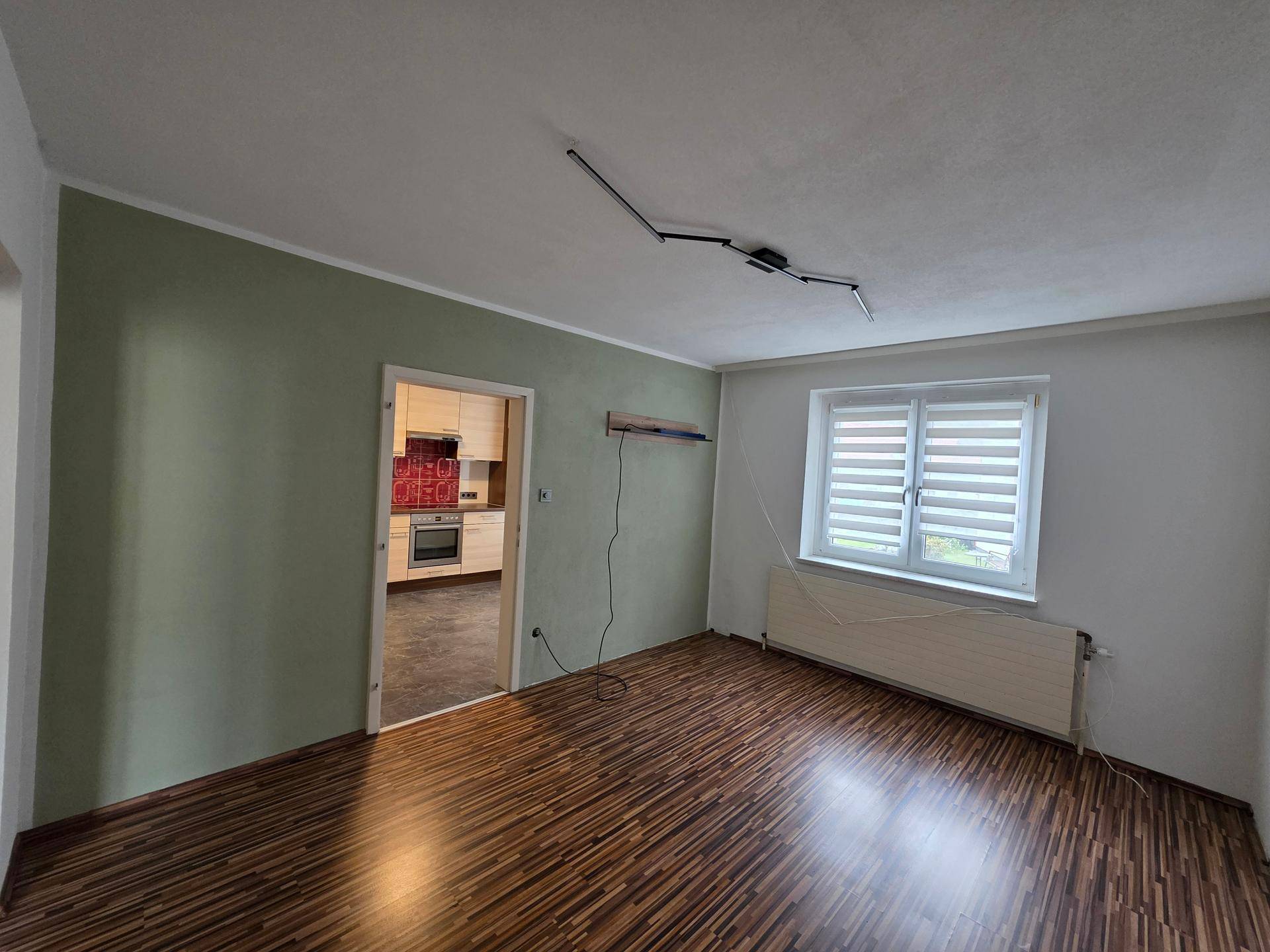 Leeres Wohnzimmer mit Laminatboden, grün gestrichener Wand und Fenster, mit Zugang zur Küche.