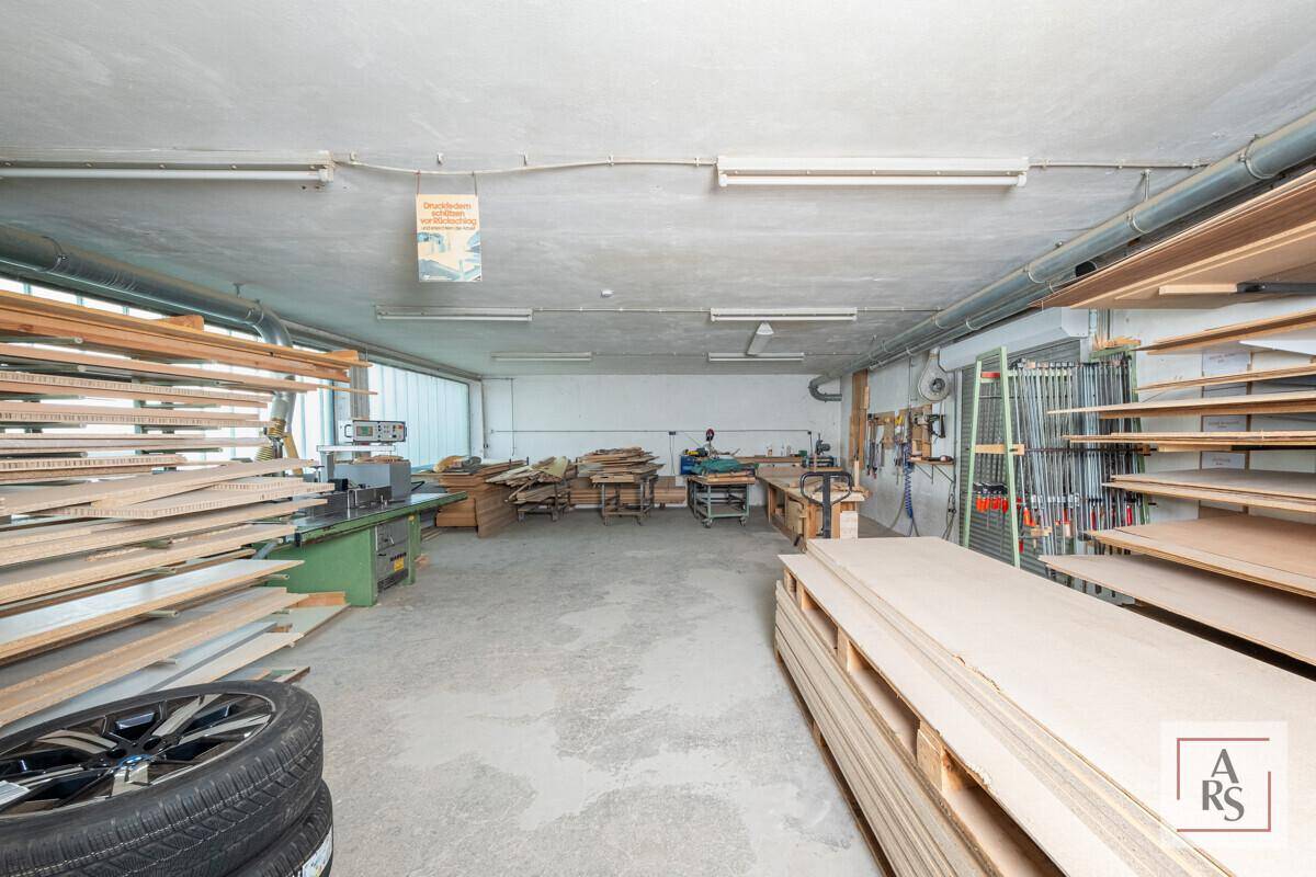 Werkstatt mit Holzbearbeitungsmaschinen, Holzstapeln und einem robusten Betonboden unter Leuchtstoffröhrenbeleuchtung.