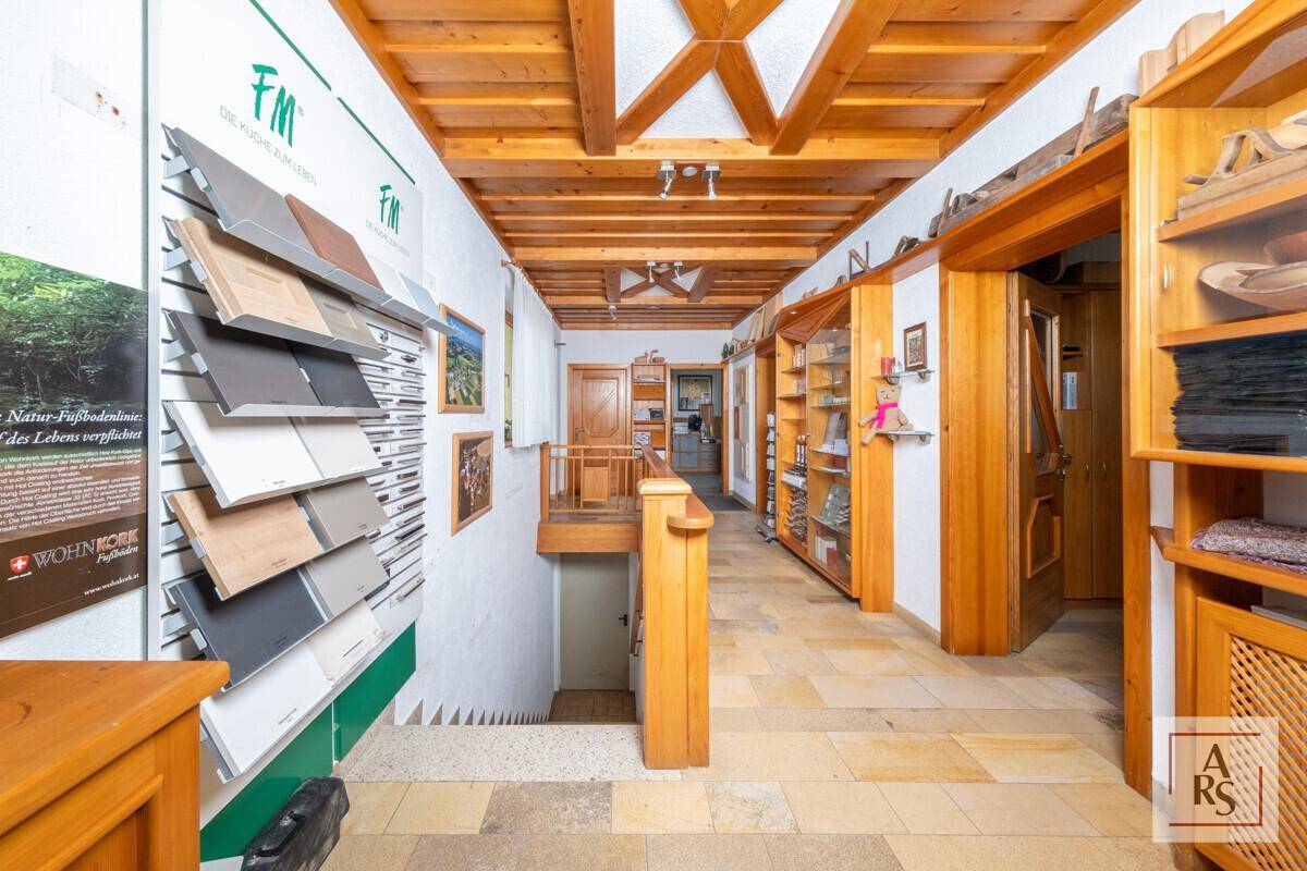 Flur mit Holzbalkendecke, Steinfliesenboden und Ausstellungsregalen für Bodenbeläge, führt zu einer Treppe.