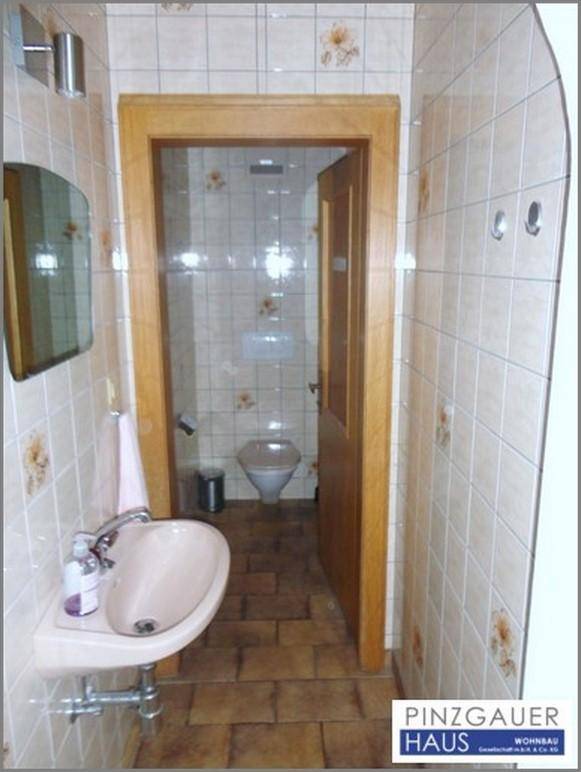 Kleines Gäste-WC mit beigefarbenen Fliesen, einem Waschbecken und einer Holztür, die zur Toilette führt.