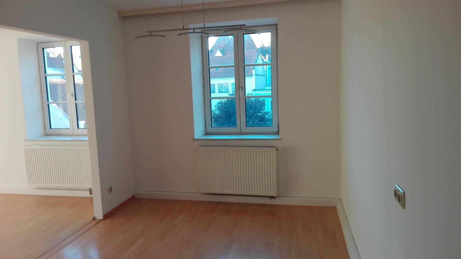 Heller Raum mit Holzboden und einem Fenster, das den Blick auf die umliegenden Gebäude ermöglicht.