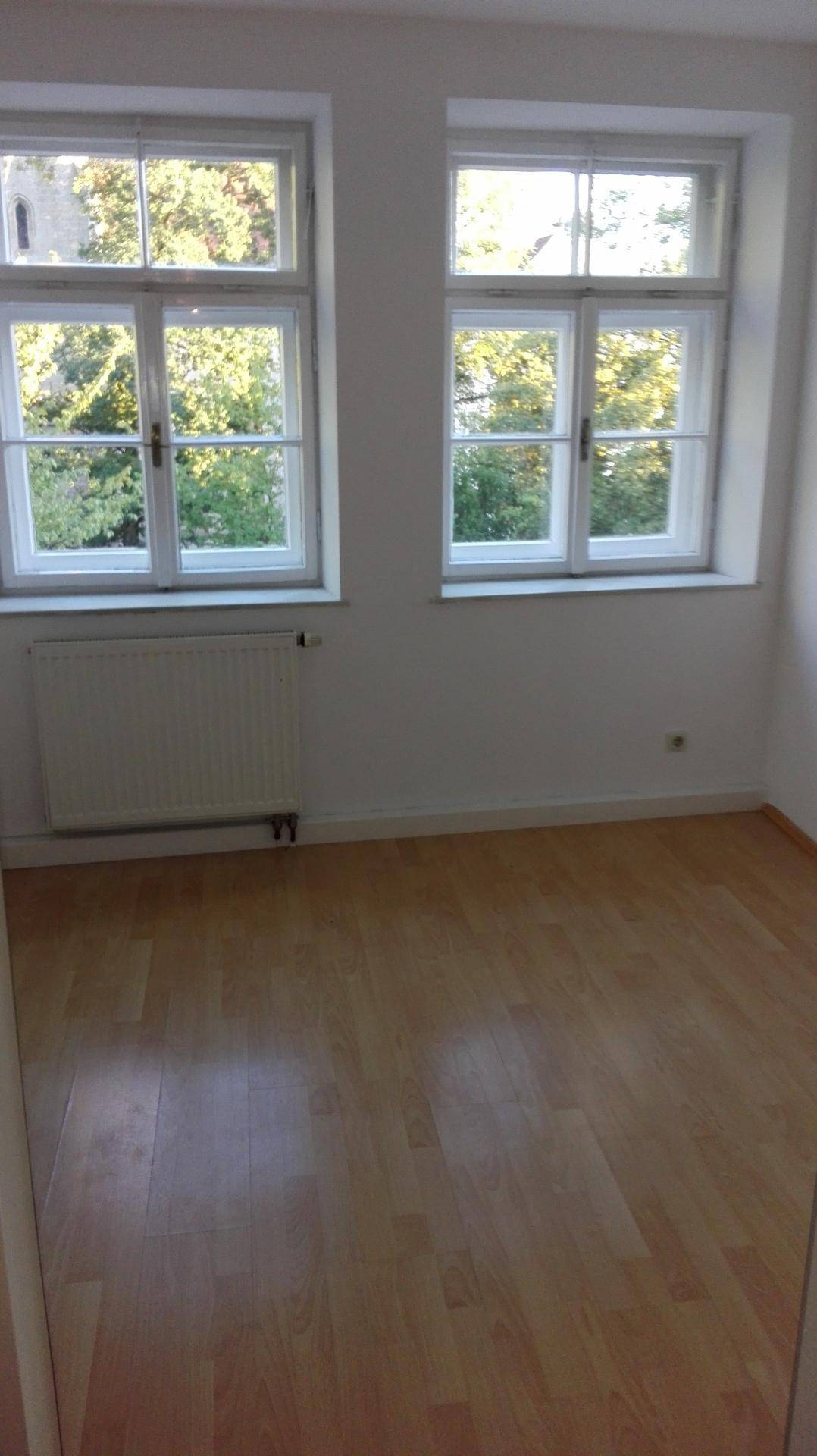 Heller Raum mit Holzboden und zwei Fenstern, die einen angenehmen Ausblick ins Grüne ermöglichen.