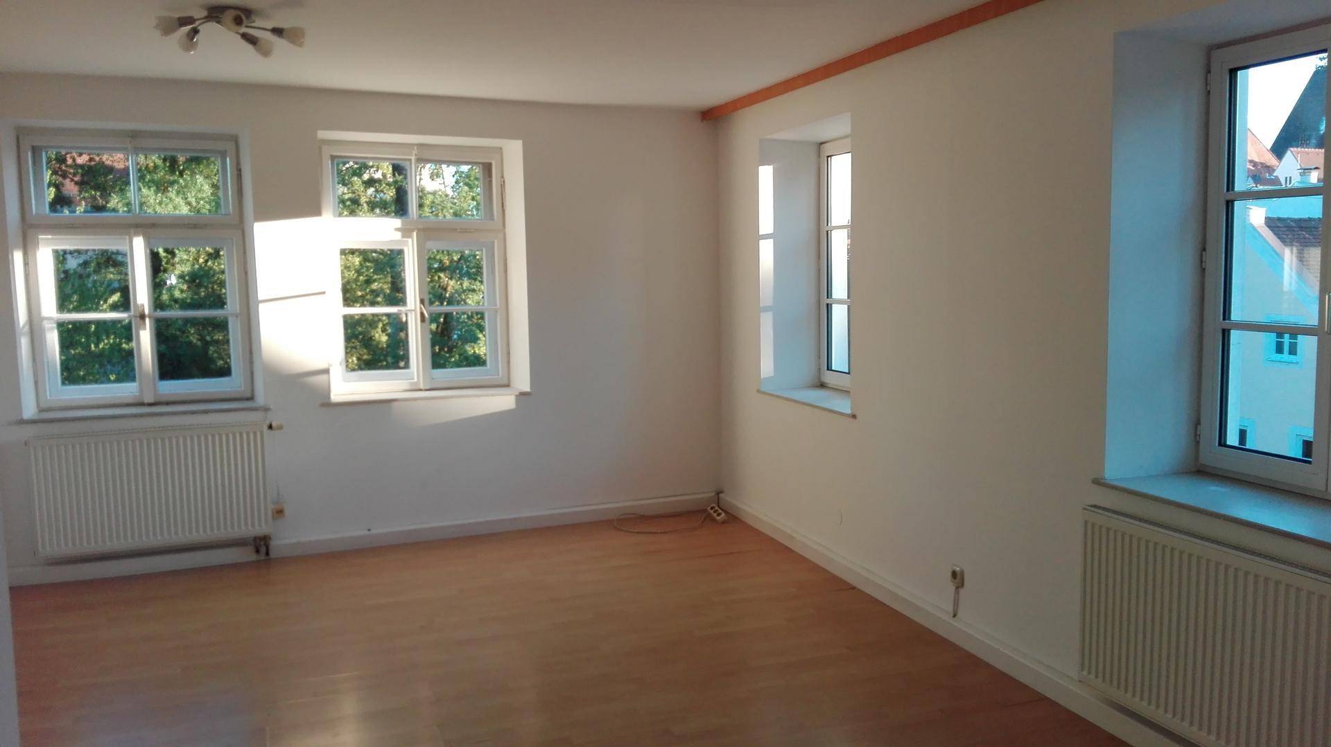 Großes, helles Zimmer mit Holzboden und mehreren Fenstern, die viel Sonnenlicht hereinlassen.
