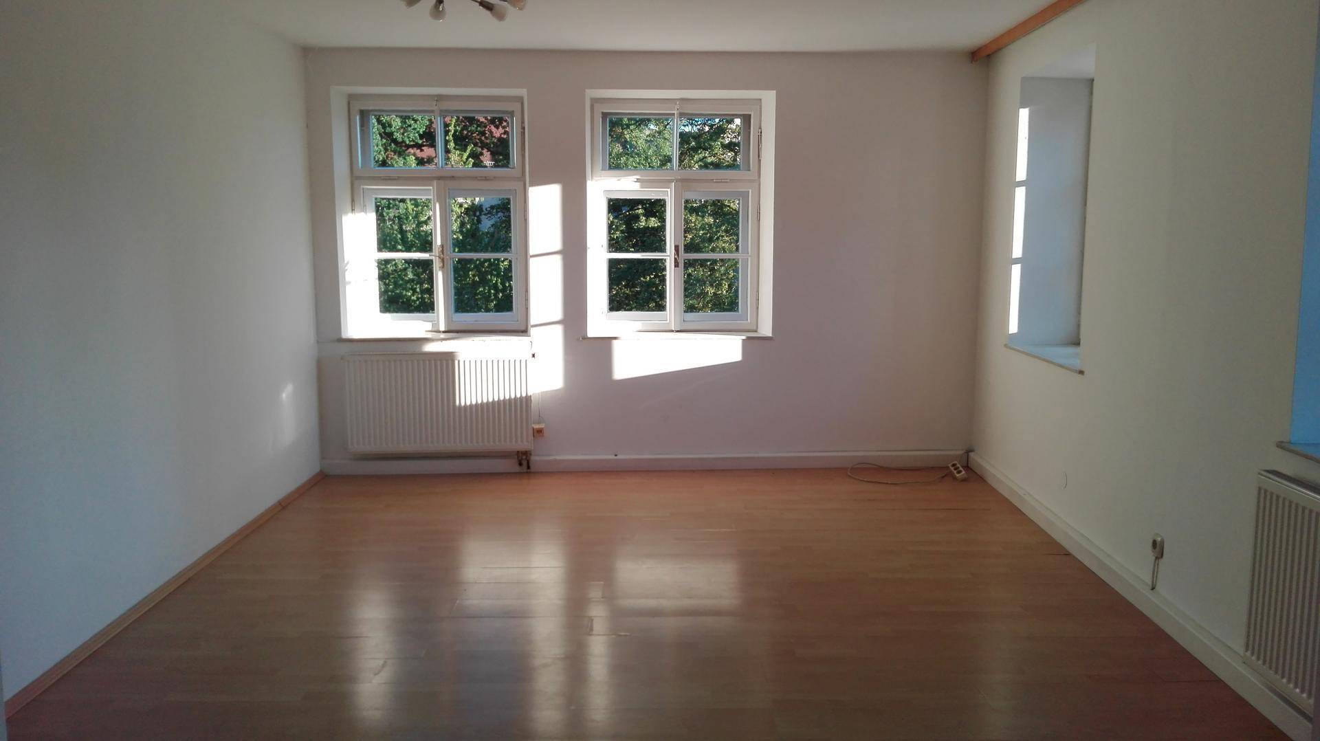 Geräumiges, sonniges Zimmer mit Holzboden und großen Fenstern, die einen grünen Ausblick bieten.