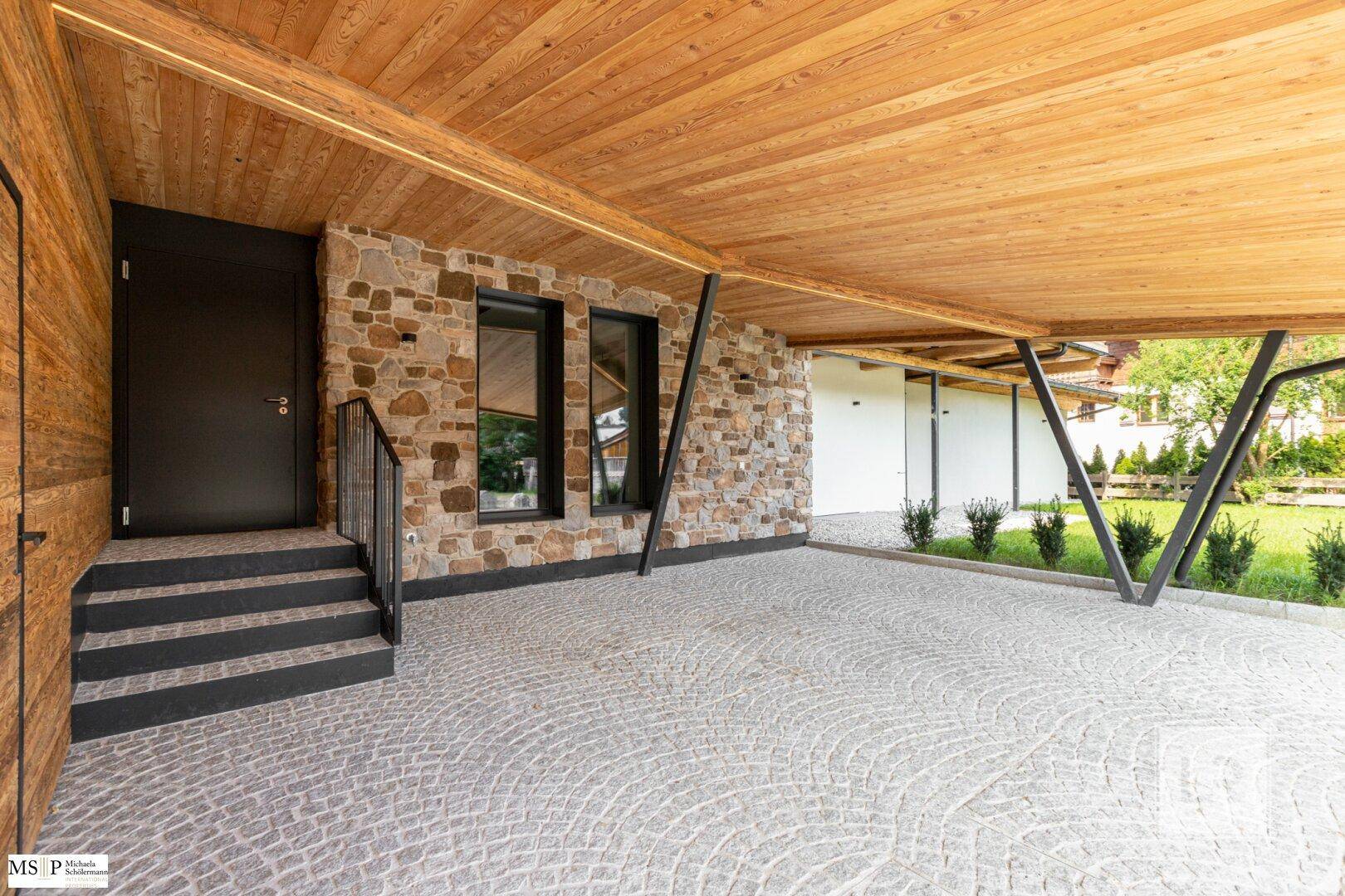 Überdachter Parkplatz mit Natursteinwand und Holzdecke, bietet Schutz für Fahrzeuge.