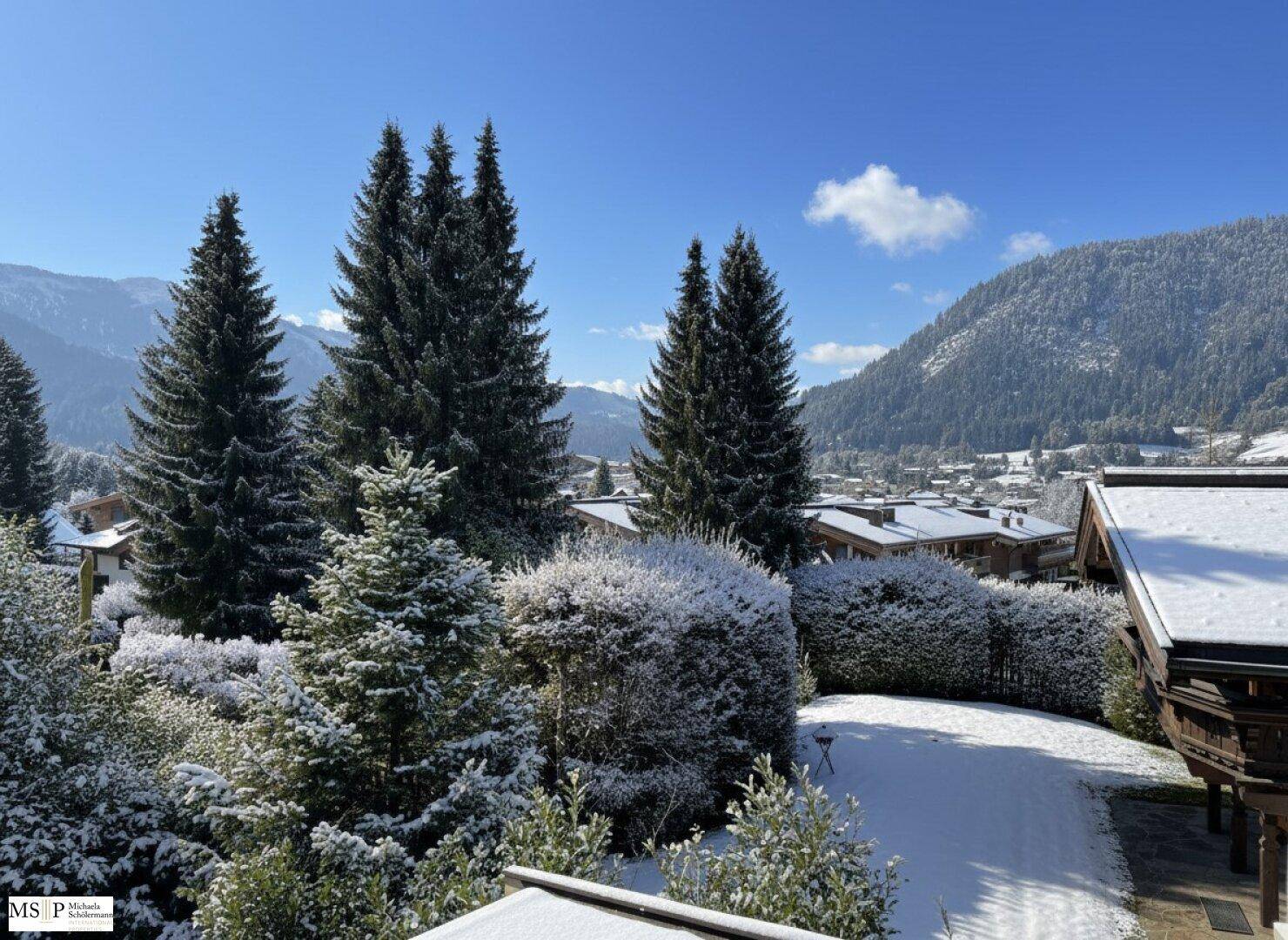 Winterlicher Ausblick auf verschneite Bäume, Dächer und Berge unter einem klaren blauen Himmel.