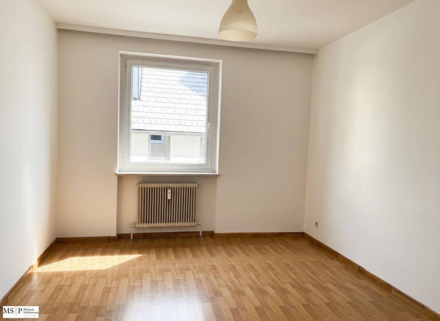 Leeres Schlafzimmer mit Parkettboden, großem Fenster und viel Tageslicht.