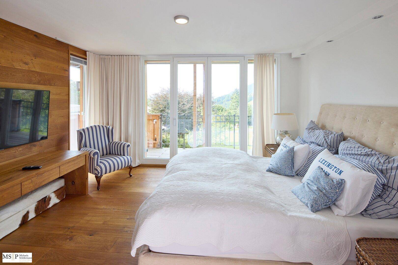 Helles Schlafzimmer mit Holzwand, Balkonzugang und Blick auf die grüne Umgebung.