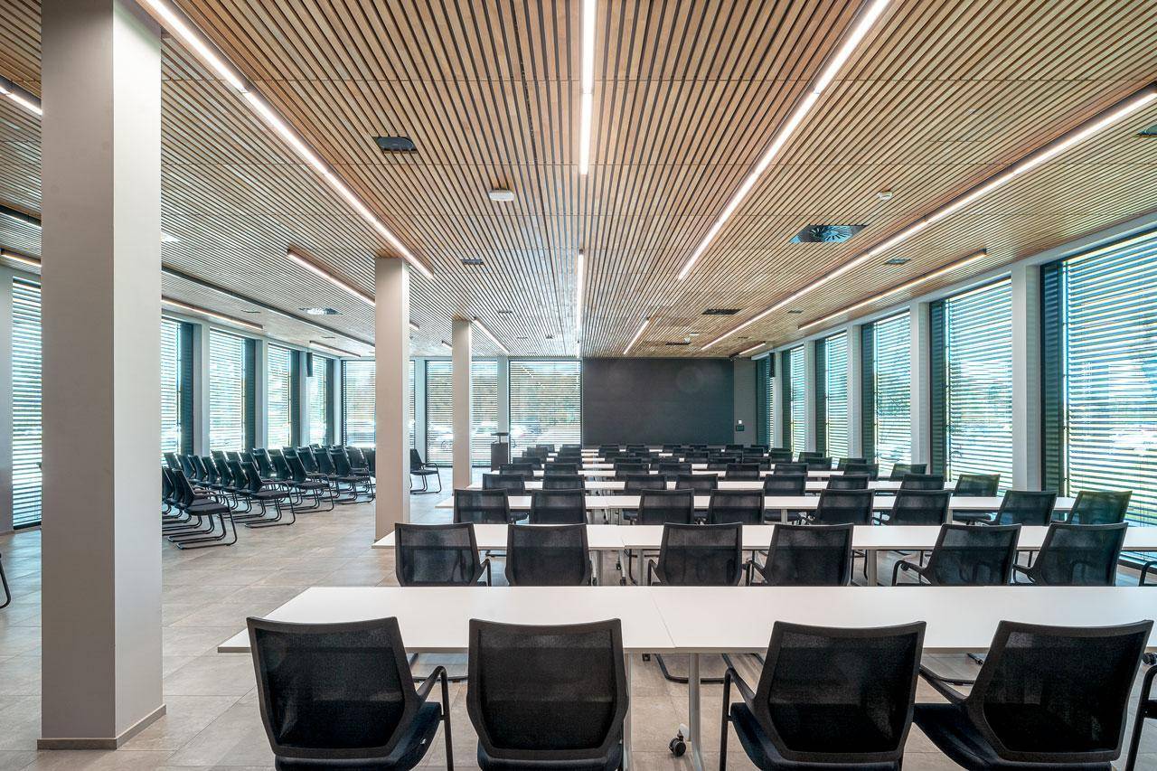 Großer Konferenz- oder Veranstaltungsraum mit Reihen von Tischen und Stühlen, Holzakzenten an der Decke und großen Fenstern.