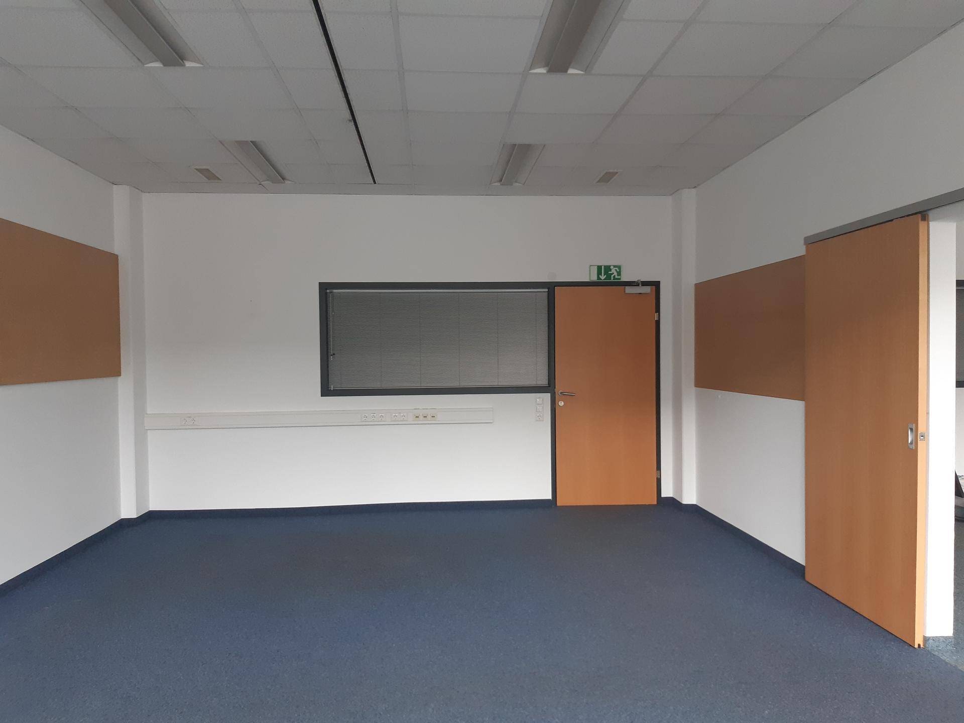 Leerer Büroraum mit blauem Teppichboden, weißen Wänden und einer Holztür.