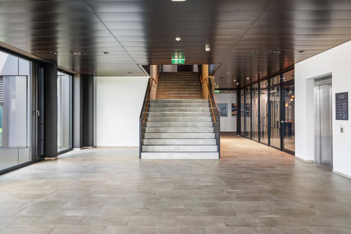 Geräumiger Flur mit Treppe, Aufzug und großen Fenstern, die viel Licht hereinlassen.