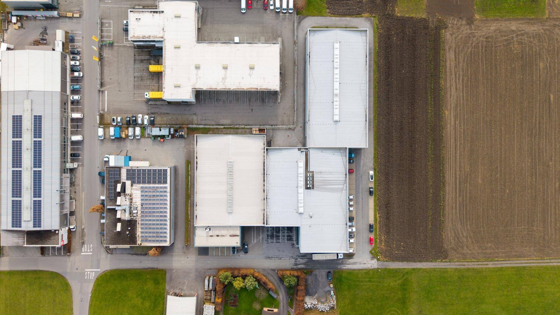 Senkrechte Luftaufnahme eines Gewerbegebiets mit verschiedenen Hallen, Parkplätzen und angrenzenden Ackerflächen.