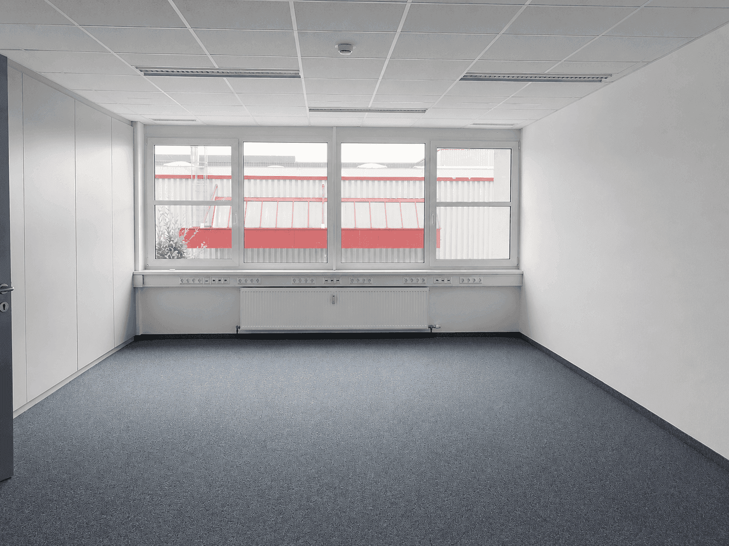 Helles Büro mit Teppichboden, breiter Fensterfront, Heizkörpern und funktionaler Deckenbeleuchtung.