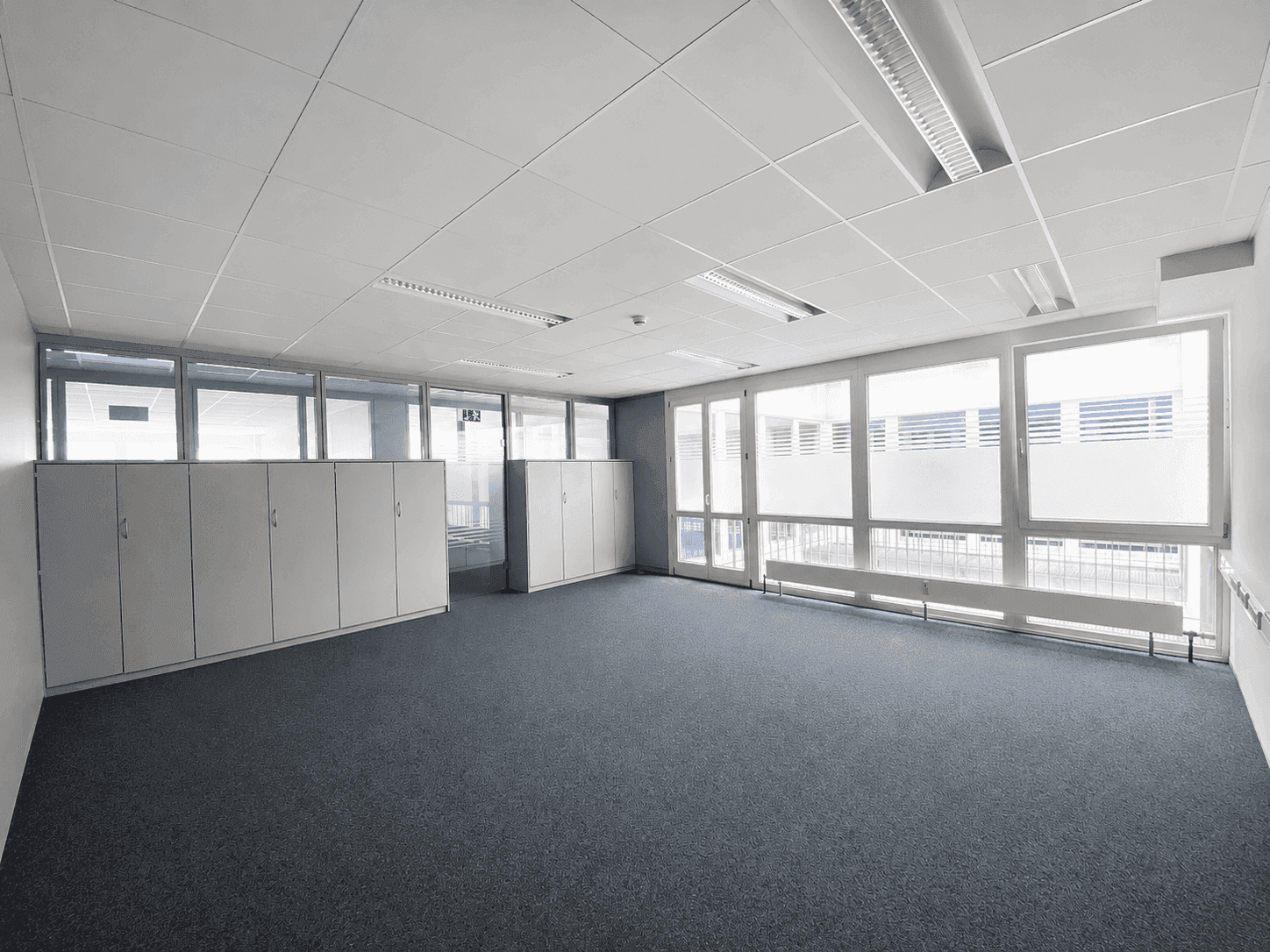Geräumiges Büro mit Teppichboden, großen Fenstern, integrierten Schränken und Deckenbeleuchtung.