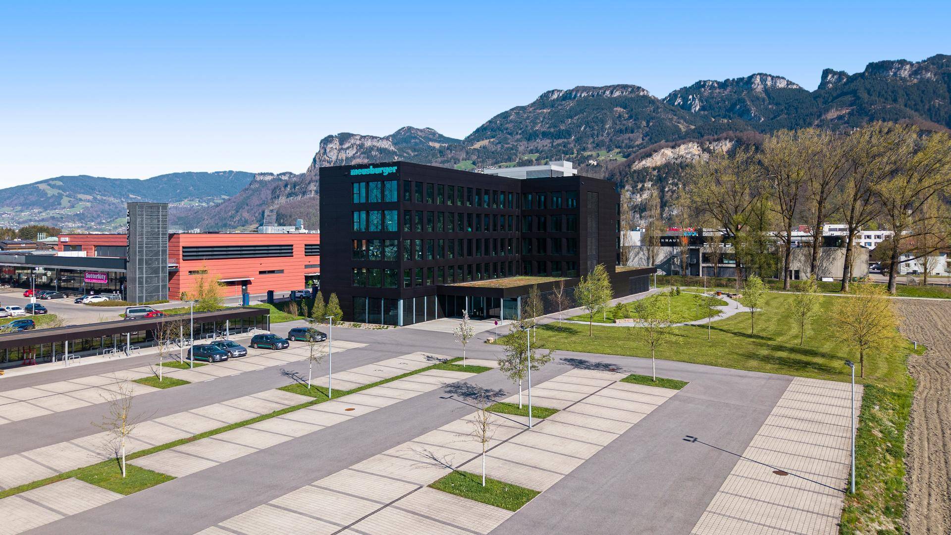 Luftaufnahme des modernen Bürogebäudes mit Parkplätzen und umliegender grüner Landschaft und Bergen.