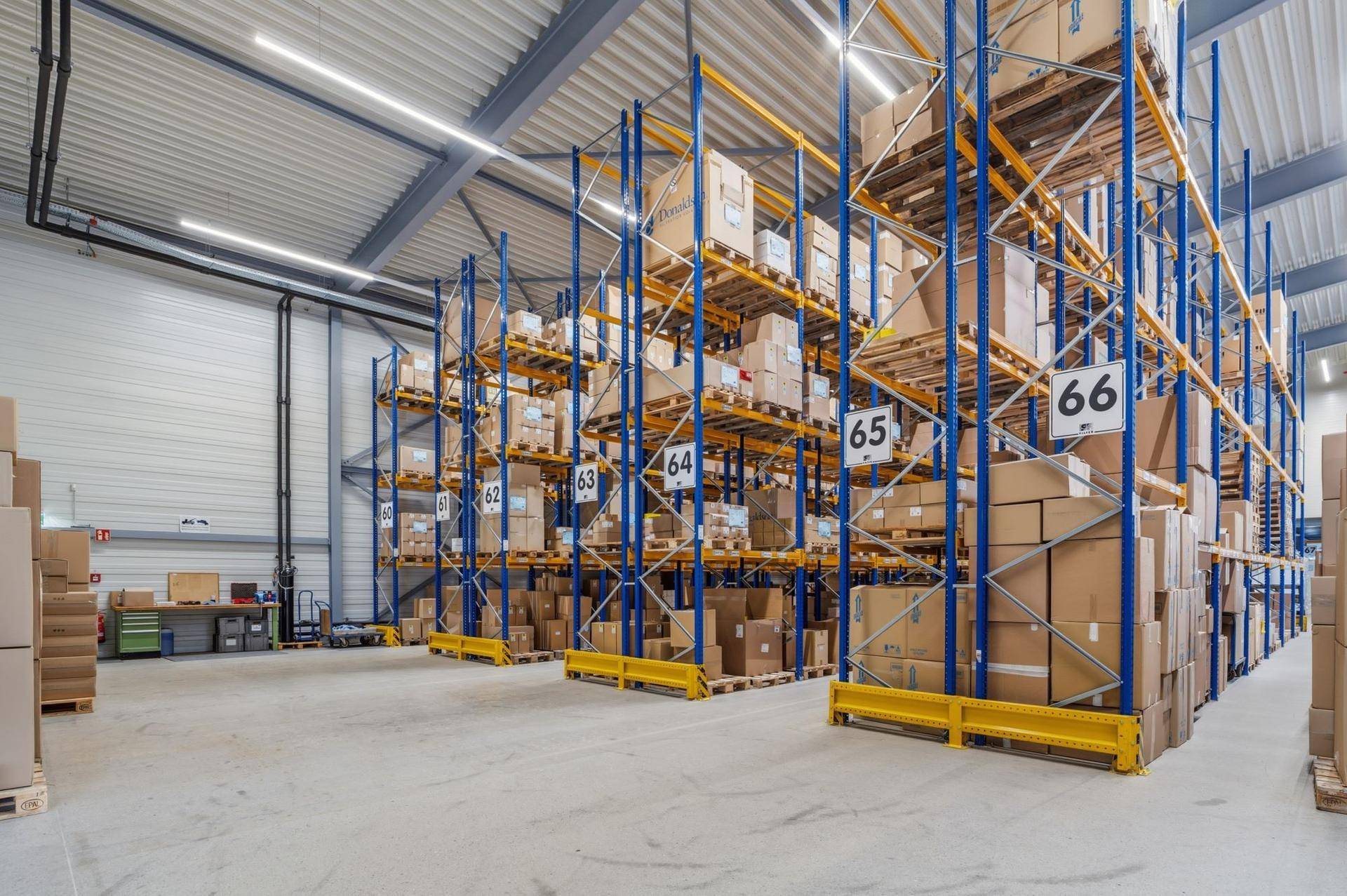 Helle Lagerhalle mit blauen und gelben Hochregalen, gefüllt mit verpackten Waren und breiten Gängen.