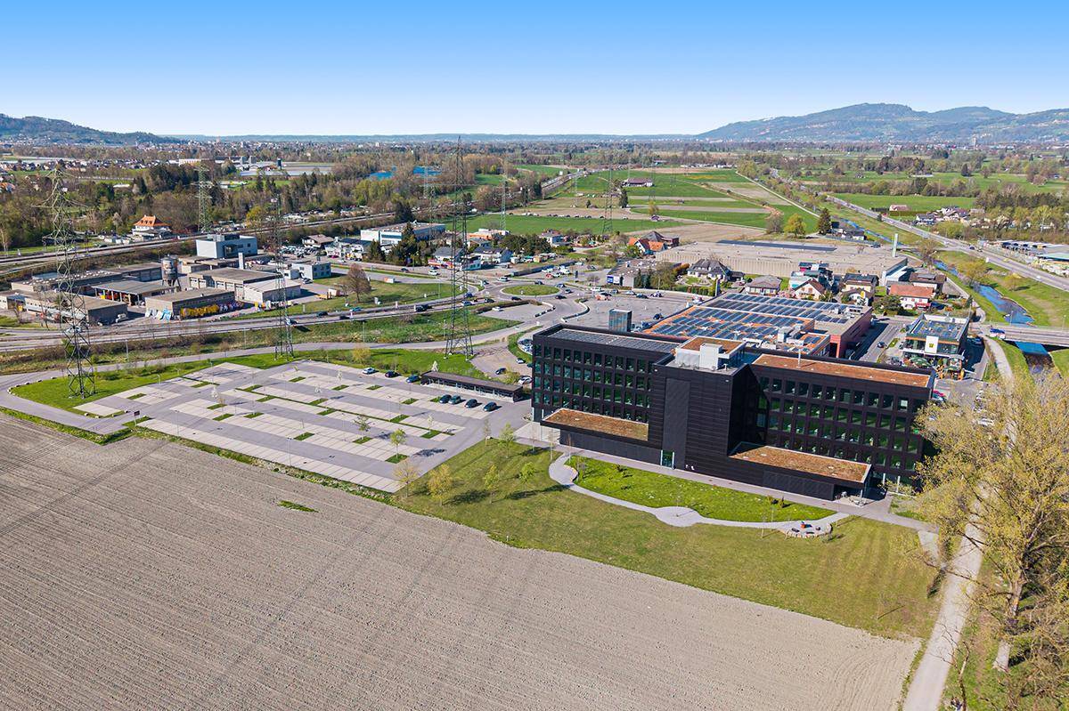 Luftaufnahme des modernen Bürogebäudes mit umliegenden Feldern und Parkplätzen unter blauem Himmel.
