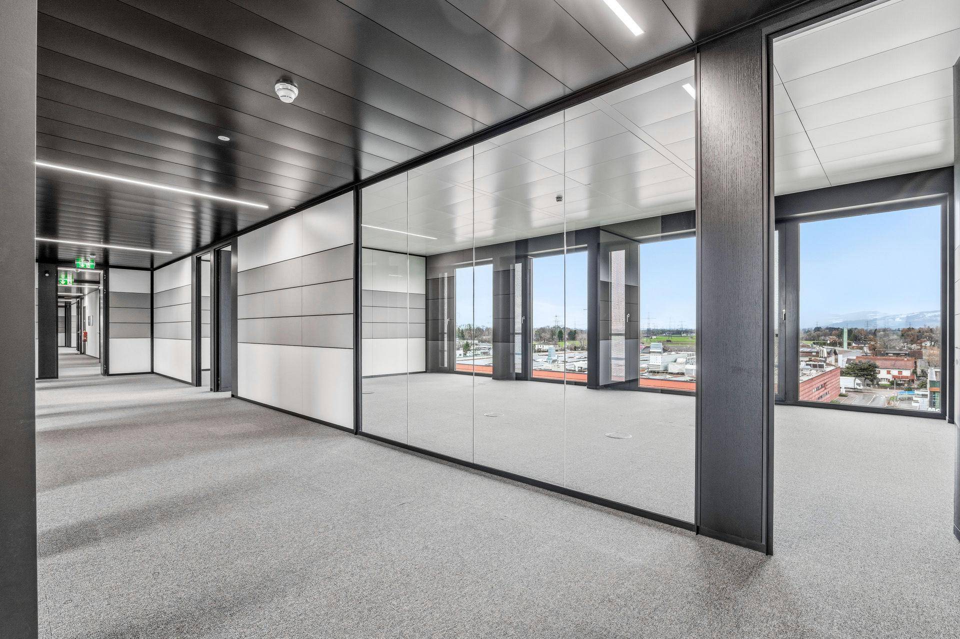 Moderner Bürogang mit Glaswänden und Teppichboden, der zu hellen Büroräumen mit weitem Ausblick führt.