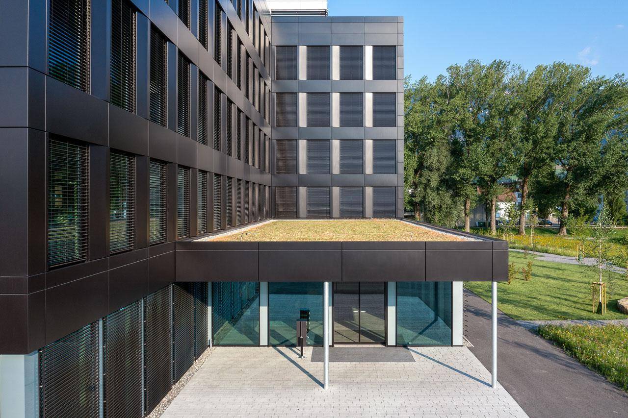 Moderner Eingangsbereich eines Bürogebäudes mit Glasfassade und begrüntem Dach, umgeben von Bäumen.