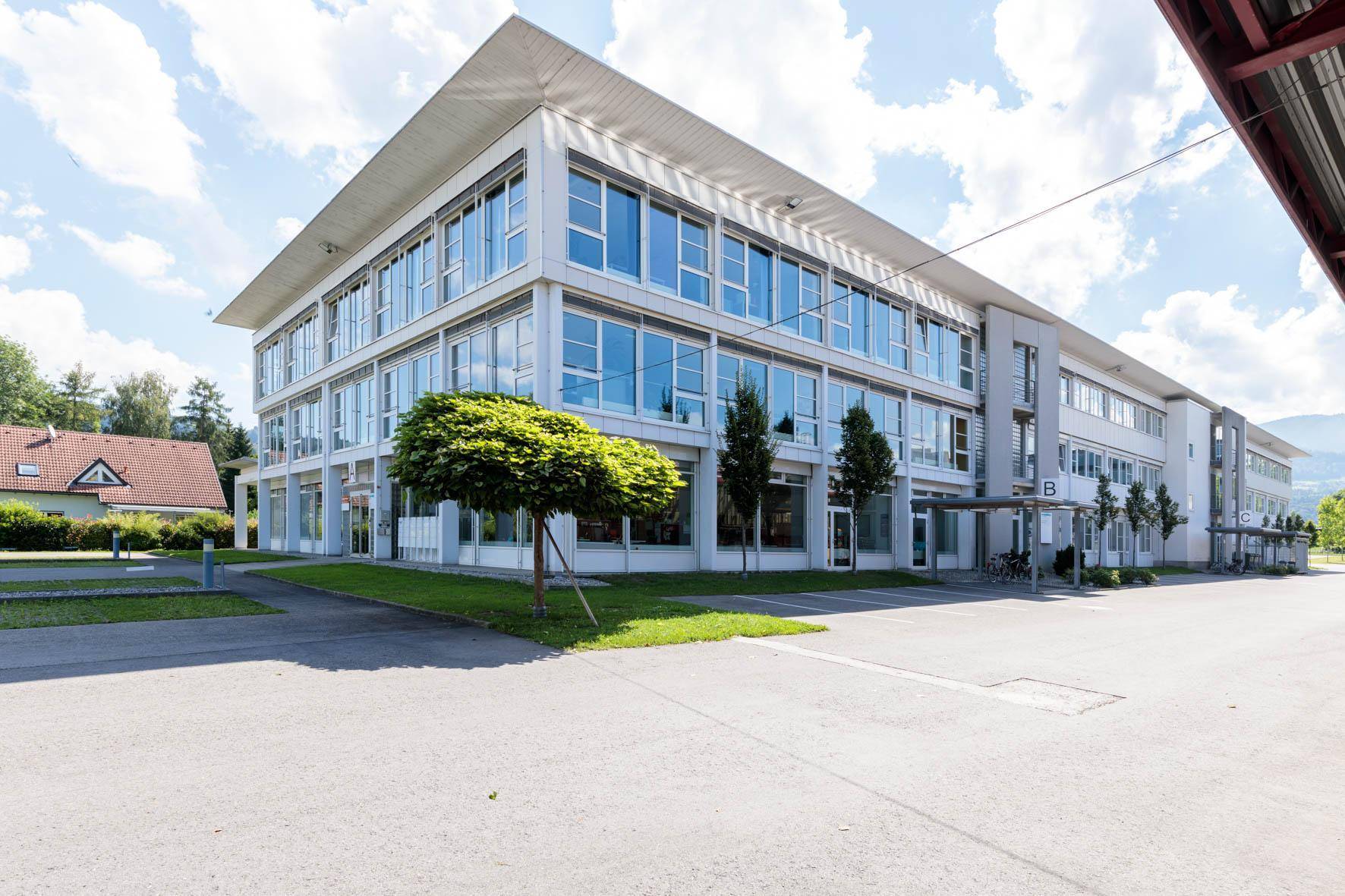 Modernes Bürogebäude mit großen Fenstern, umgeben von Grünflächen und einem Parkplatz, mit Bergblick.