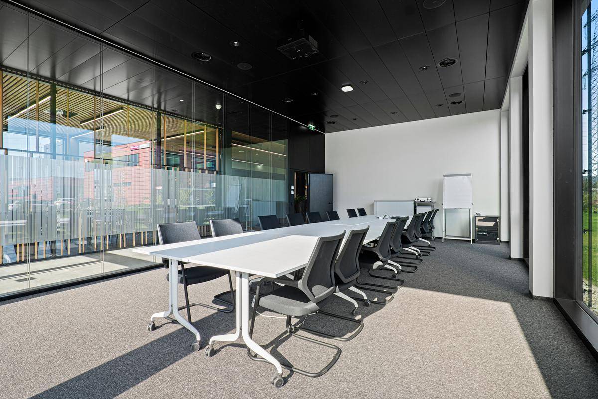 Moderner Konferenzraum mit langem Tisch, Bürostühlen und großen Fenstern, die viel Tageslicht hereinlassen.