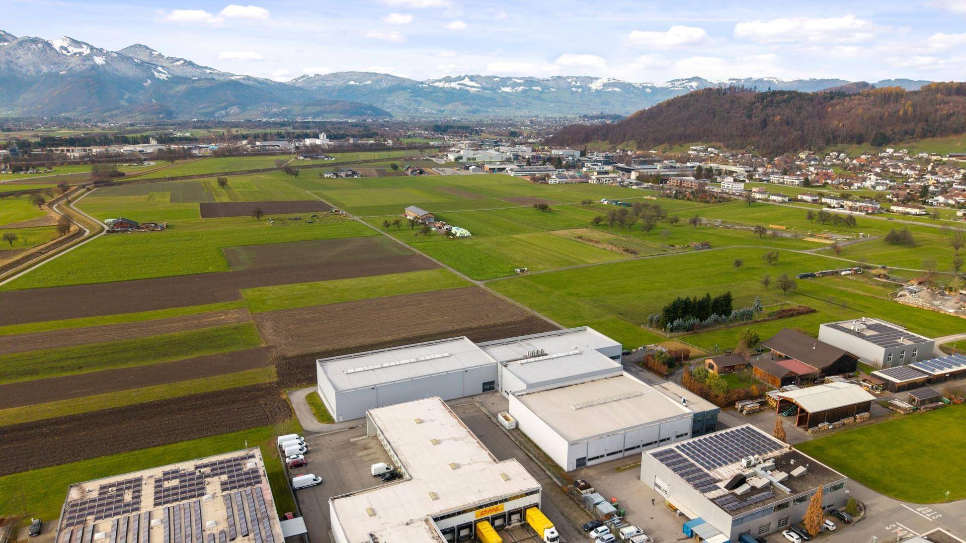 Panoramische Luftaufnahme eines Industriegebiets vor einer beeindruckenden Bergkulisse und weiten grünen Feldern.