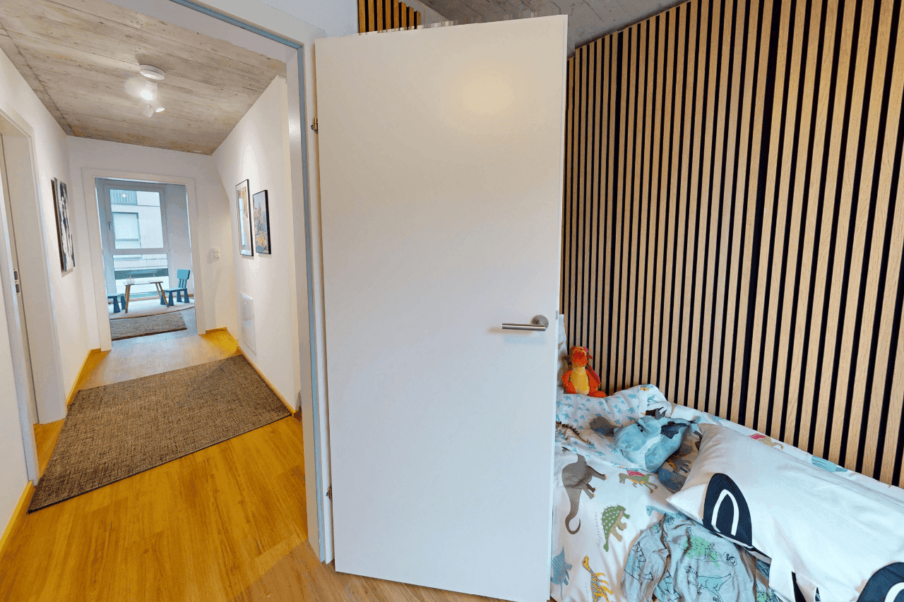 Kinderzimmer mit Holzwandverkleidung, Sichtbetondecke und Zugang zu einem hellen Flur.