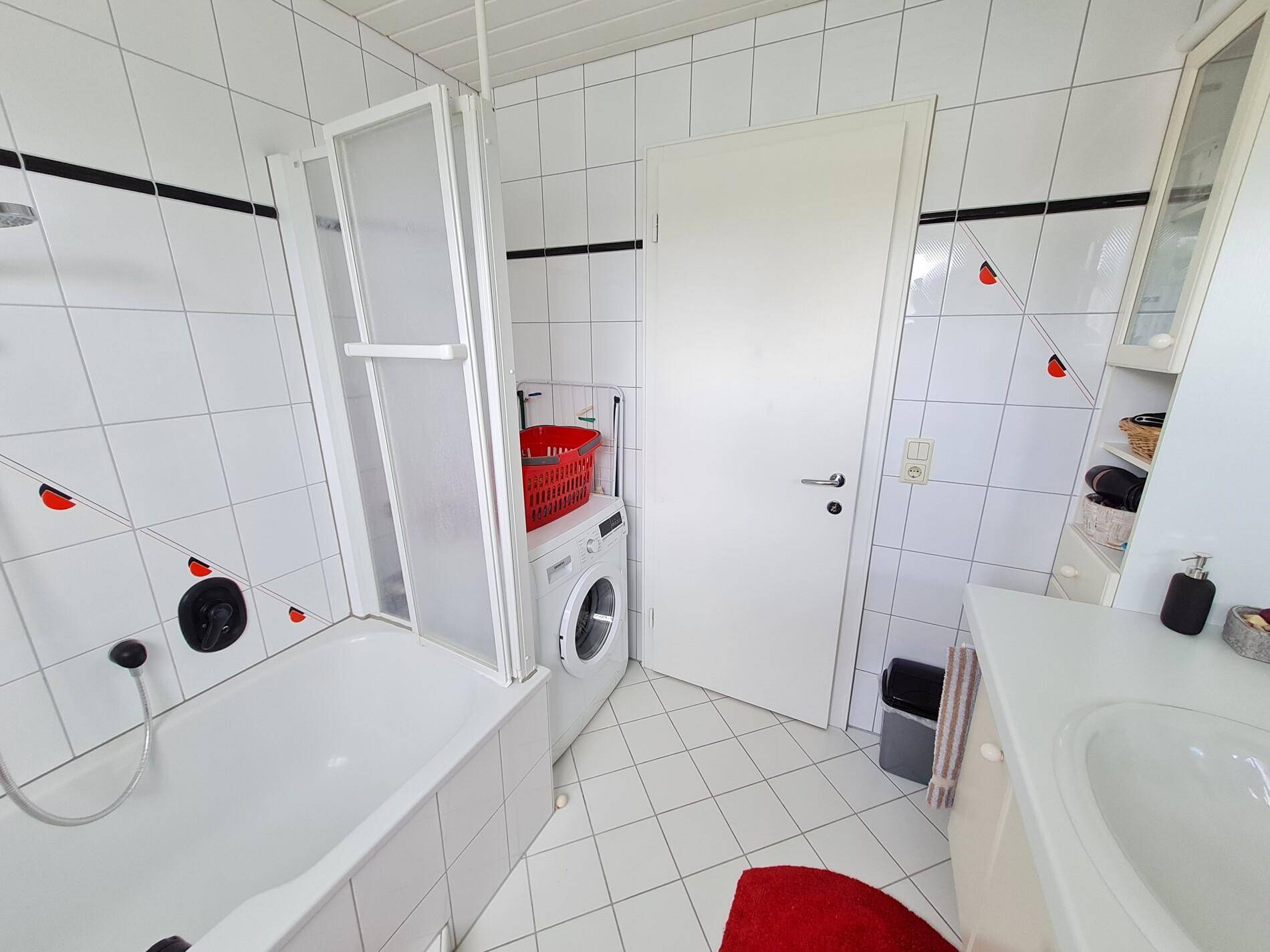 Badezimmer mit Badewanne, Duschabtrennung und integrierter Waschmaschine, hell gefliest.