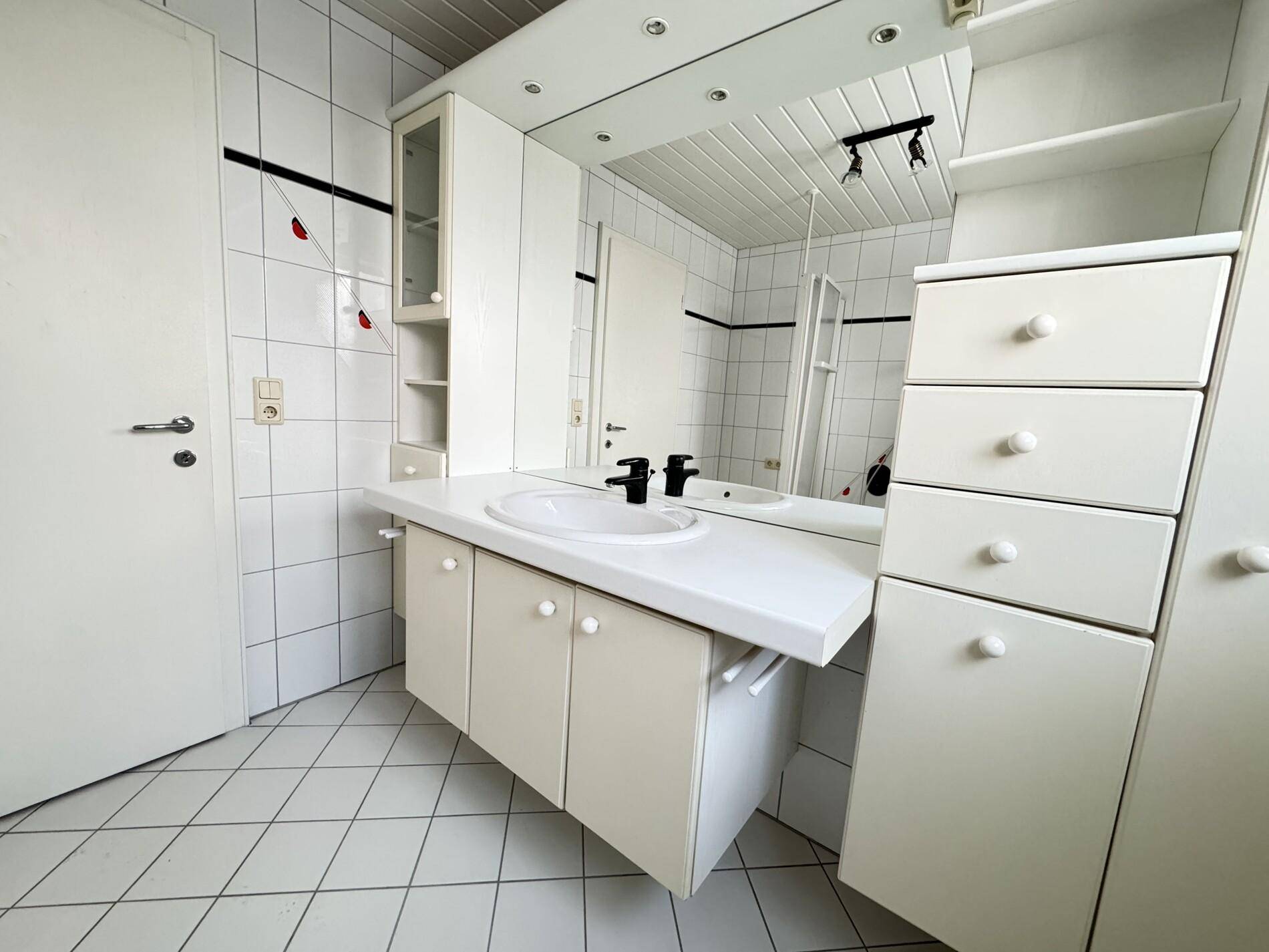 Helles Badezimmer mit weißen Fliesen, einem einzelnen Waschbecken und praktischen Spiegelschränken.