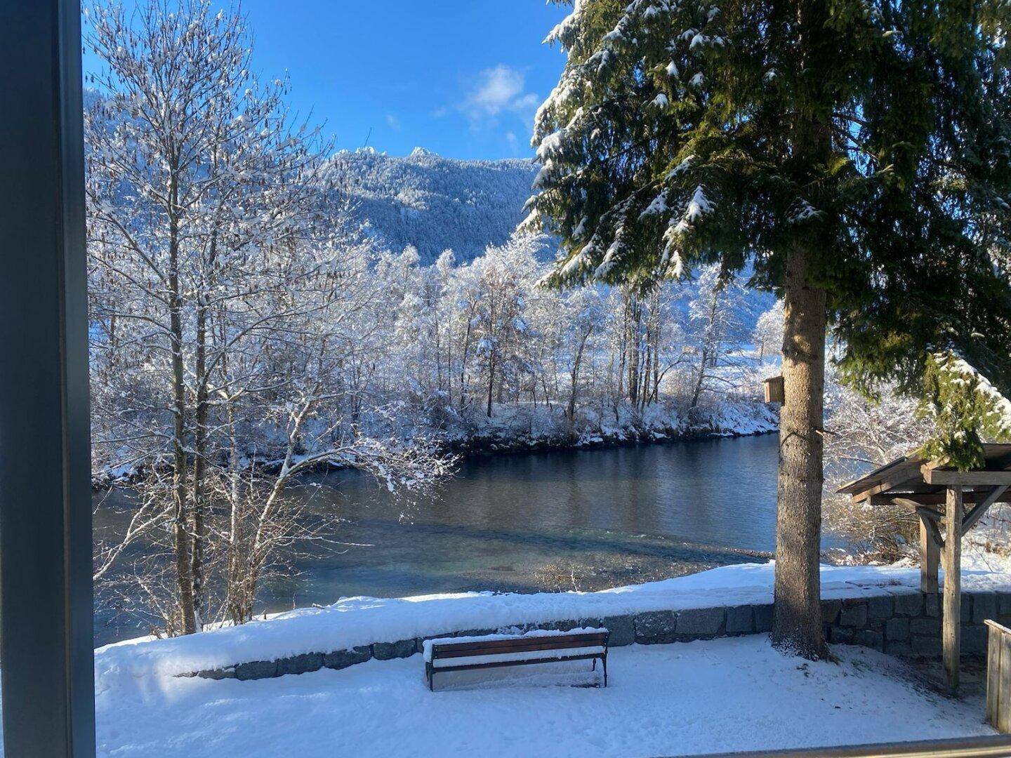 Idyllische Winterlandschaft mit schneebedeckten Bäumen, einem Fluss und Bergen im Hintergrund.