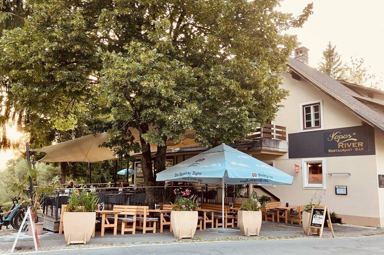 Außenansicht des Restaurants mit großer Terrasse, Sonnenschirmen und üppiger Vegetation.