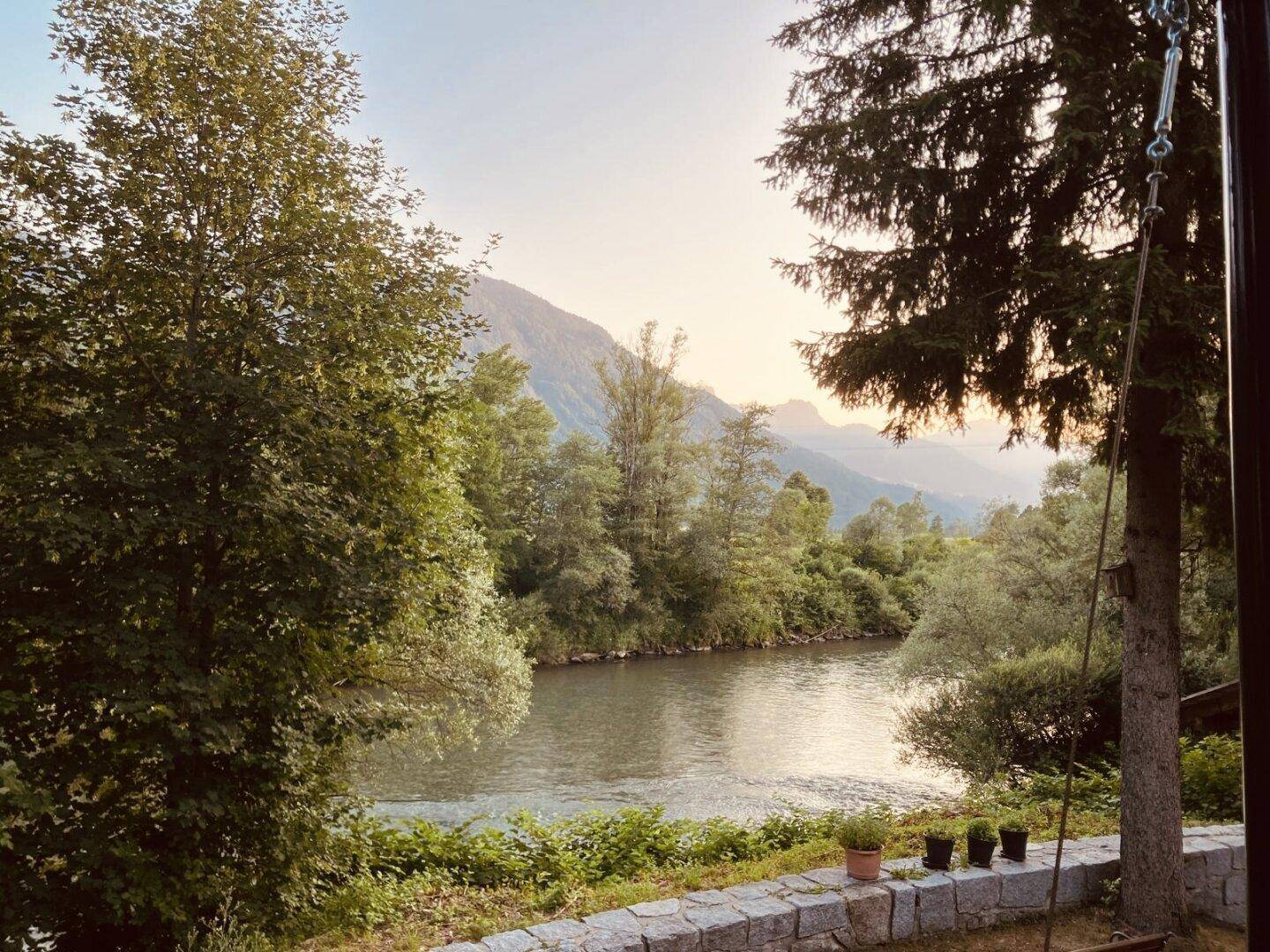 Weitläufiger Blick von der Terrasse auf den ruhigen Fluss und die majestätischen Berge bei Sonnenuntergang.
