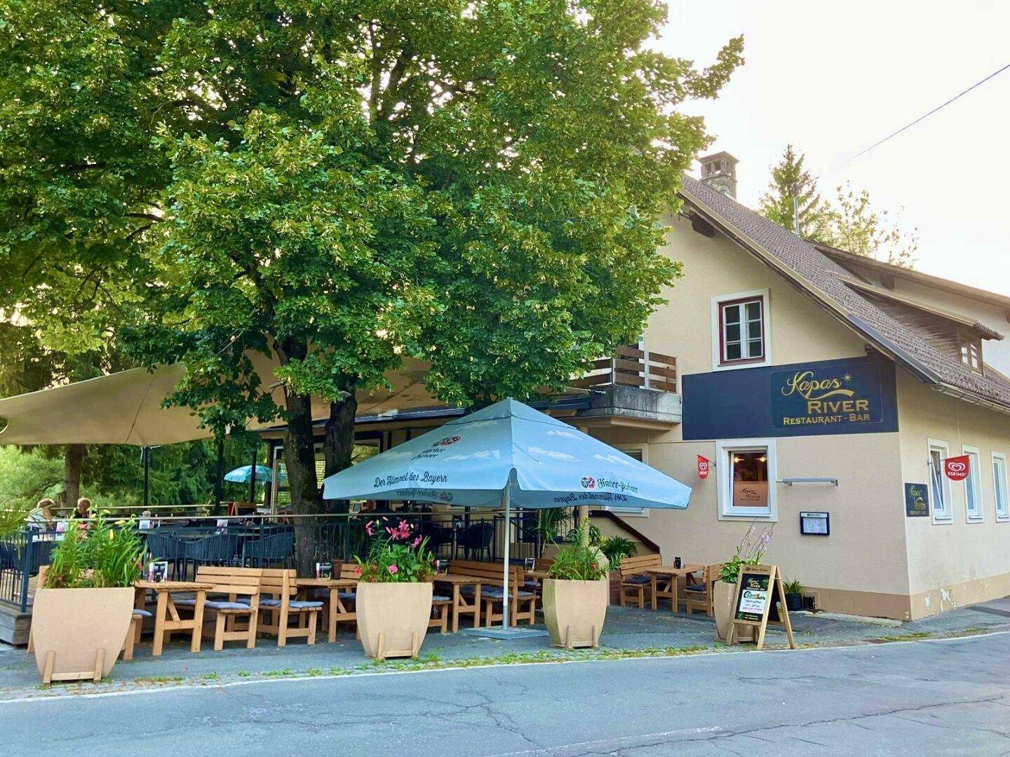 Außenansicht des Restaurants mit großer Terrasse, Sonnenschirmen und üppiger Vegetation.