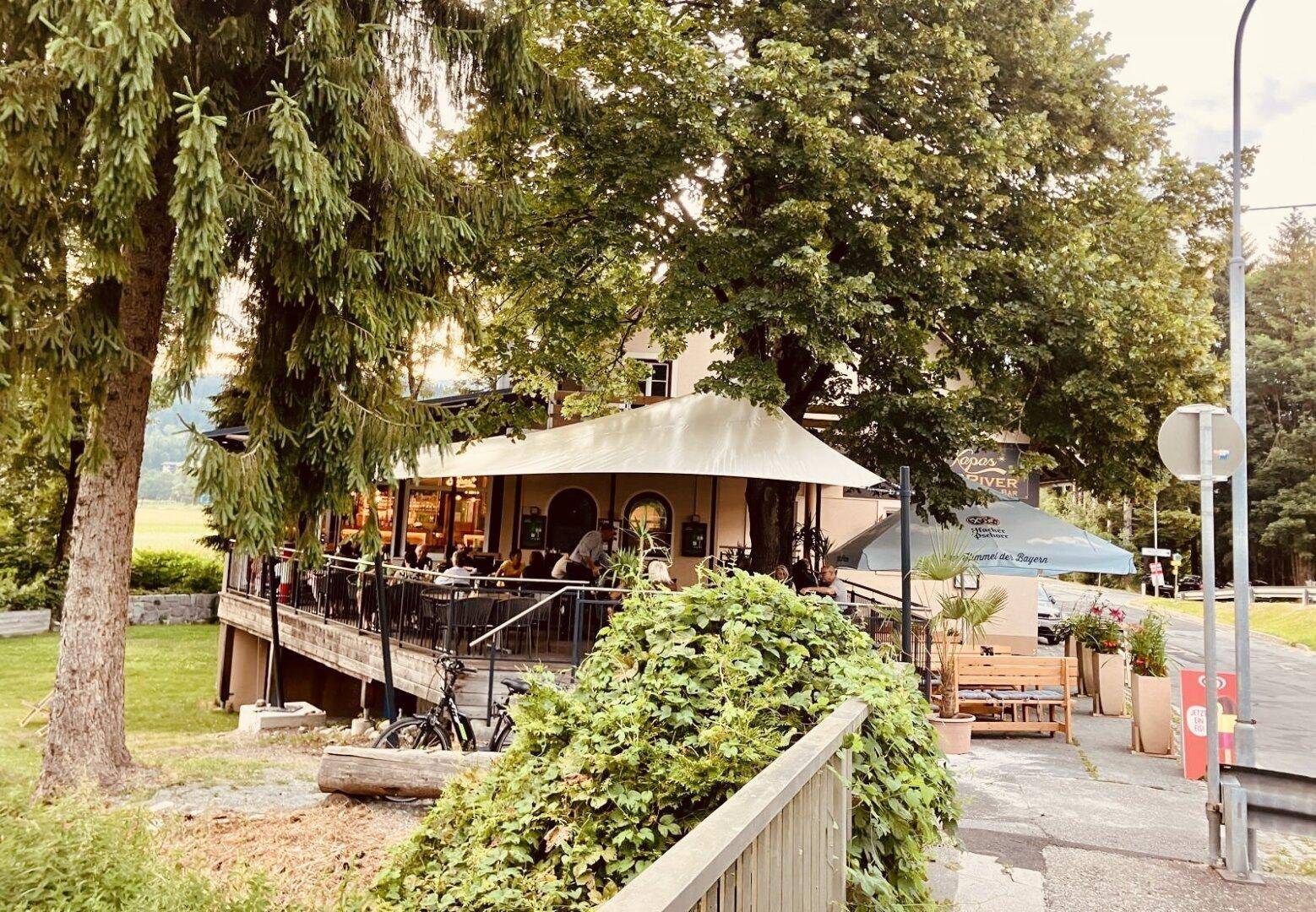Restaurantgebäude mit erhöhter Terrasse, umgeben von Bäumen und grüner Landschaft.